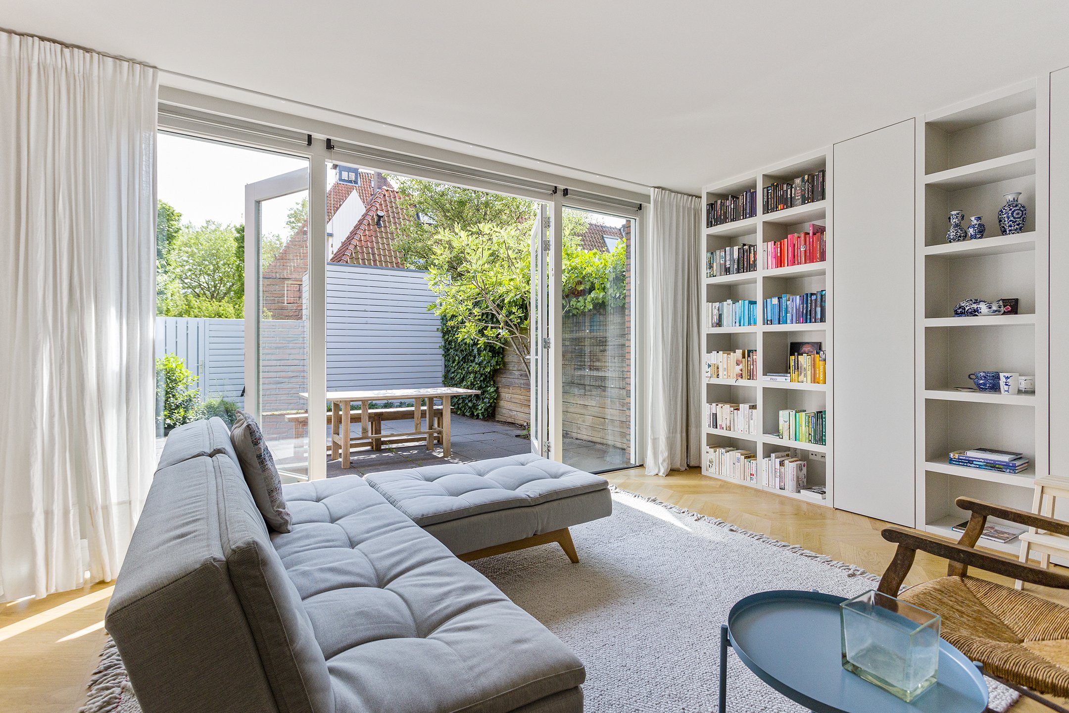 Licht woonkamer met grote glazen schuifdeuren naar tuin, gestoffeerde bank, witte boekenplanken met gekleurde boeken en décor, houten vloer, ronde blauwe salontafel, stoel met houten armleuningen, beige tapijt.