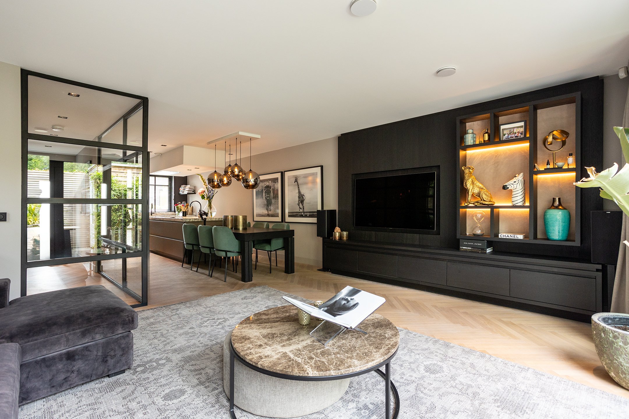 Moderne woonkamer met zwarte wandmeubel met decoraties en een grote televisie, een ronde salontafel met een open magazine, een grijze bank en een eettafel met groene stoelen, grote ramen die veel natuurlijk licht binnenlaten en een open keuken verder