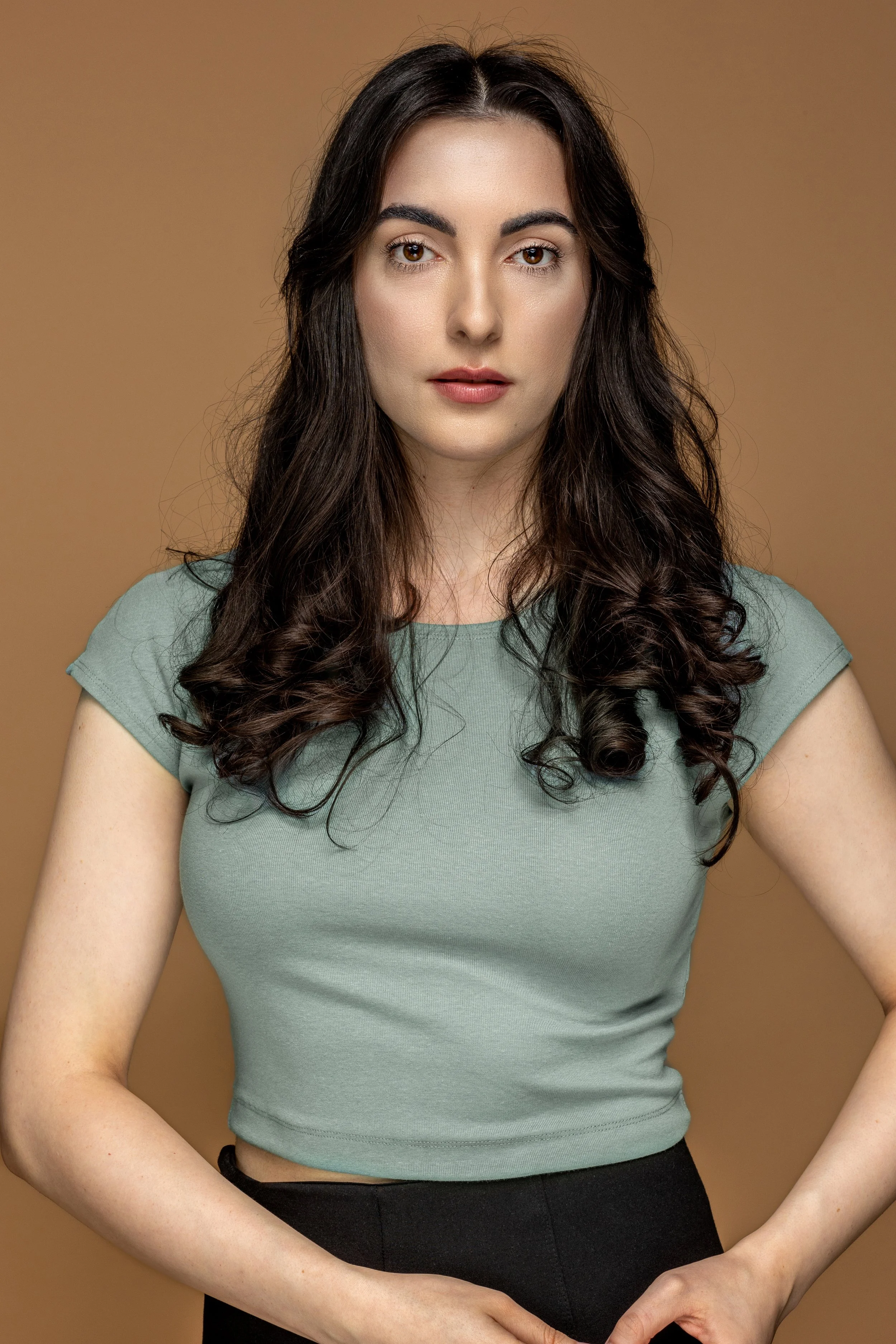 Portret van een jonge vrouw met lang, zwart, gekruld haar en lichte huid, die recht naar de camera kijkt, gekleed in een groene top en zwarte broek.