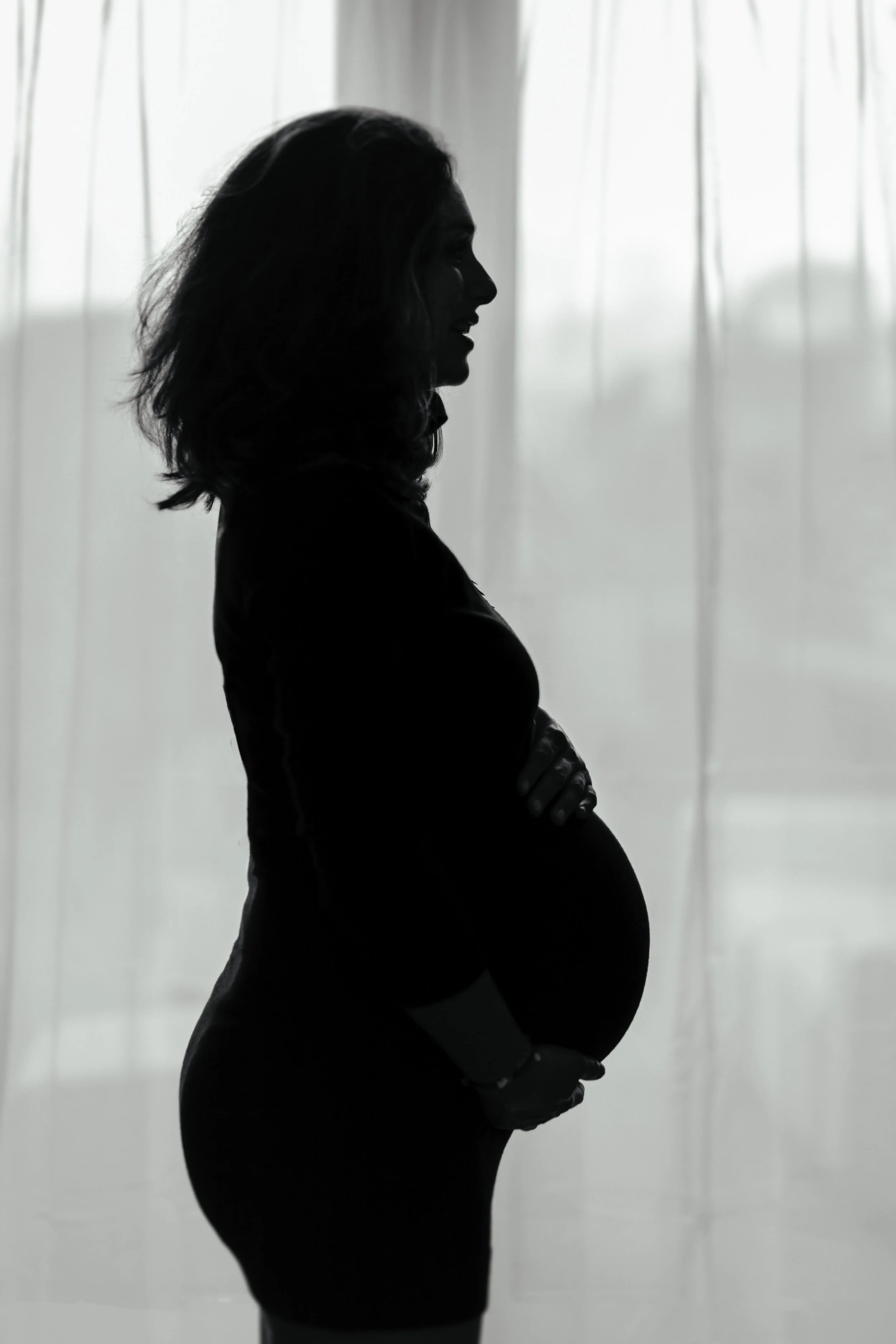 Silhouet van een zwangere vrouw die voor een raam staat met doorlatende gordijnen.