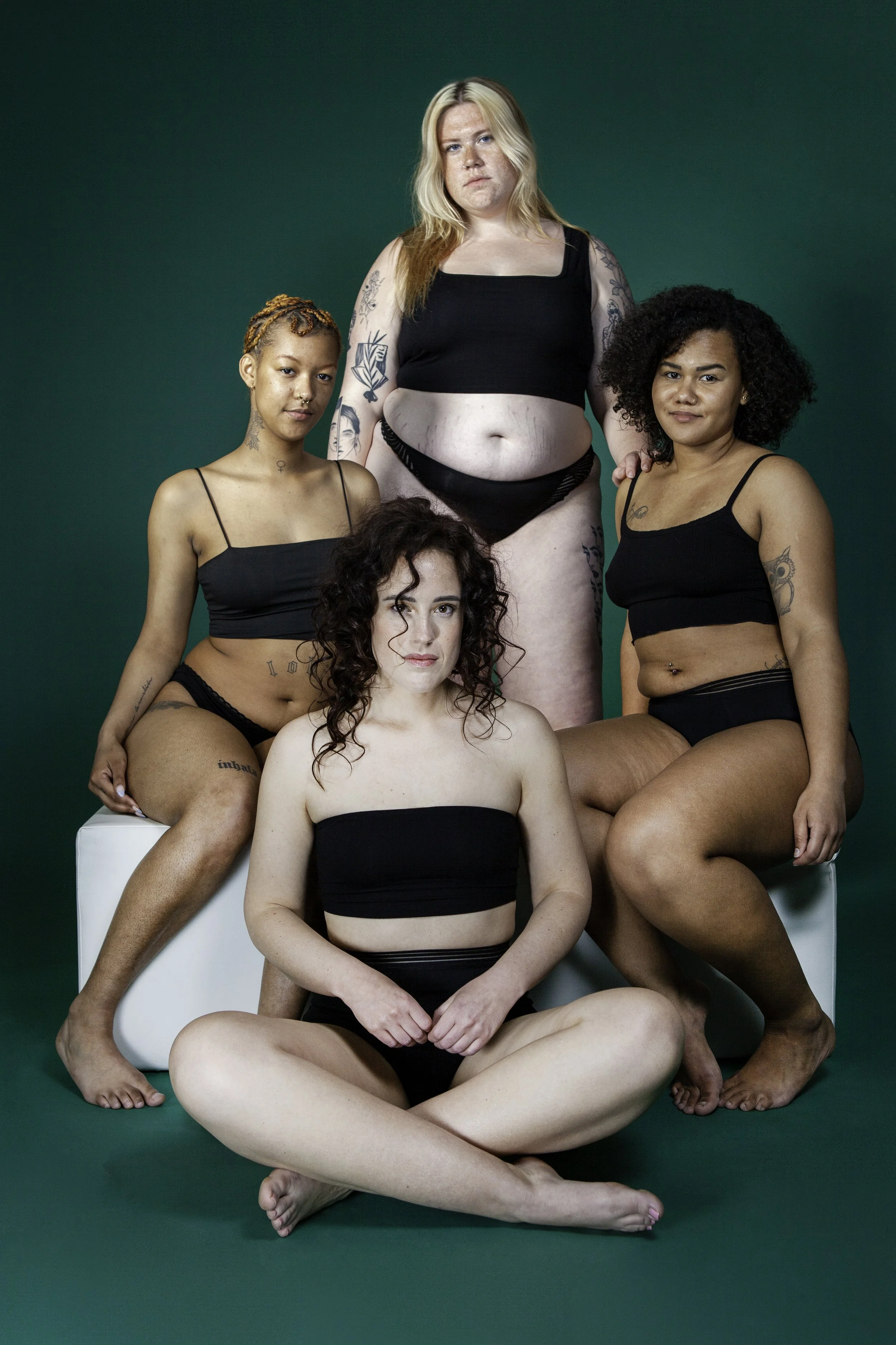 Vijf vrouwen met diverse huidtinten en lichaamsbouw, gekleed in zwart ondergoed, poseert tegen een groene achtergrond. Ze zitten en staan in een groepsfoto.