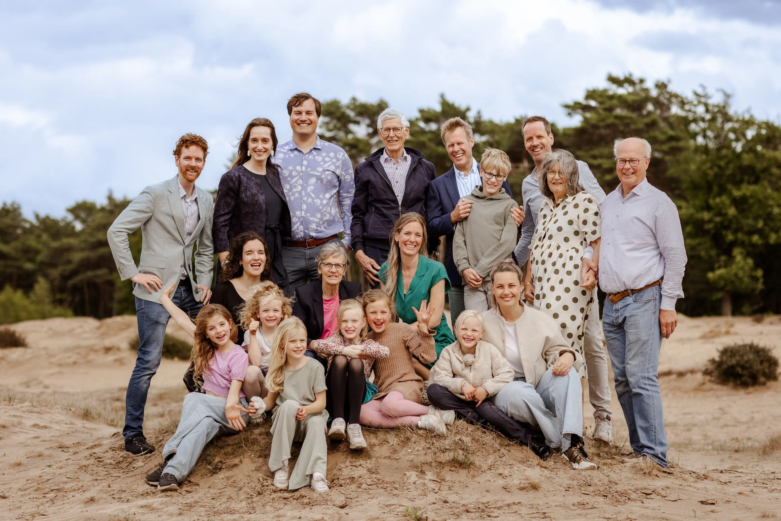 Een grote familie poseert samen buiten op een zandheuvel met bomen op de achtergrond, ze lachen en genieten van het gezelschap.