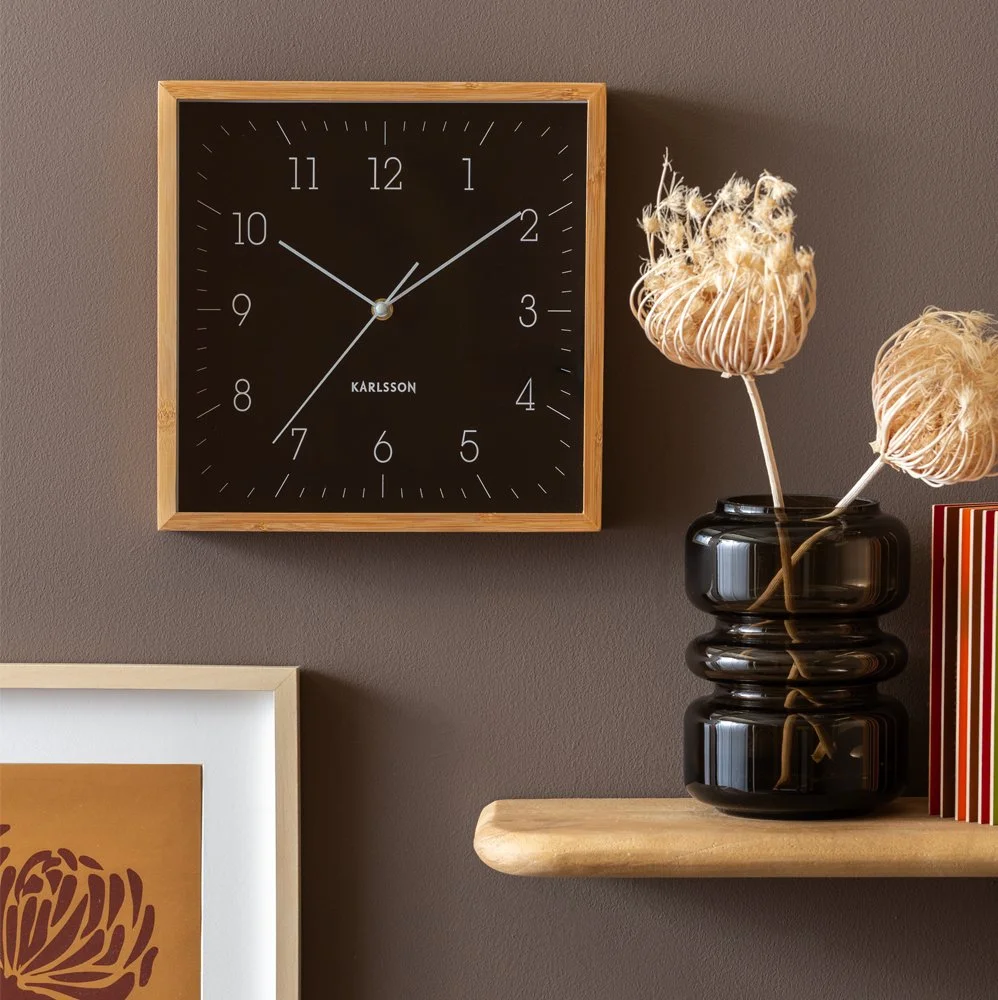 Houten wandklok met zwarte wijzerplaat en witte wijzers, geplaatst op een grijze muur, naast een zwarte glazen vaas met gedroogde bloemen en een kleurrijk boek.