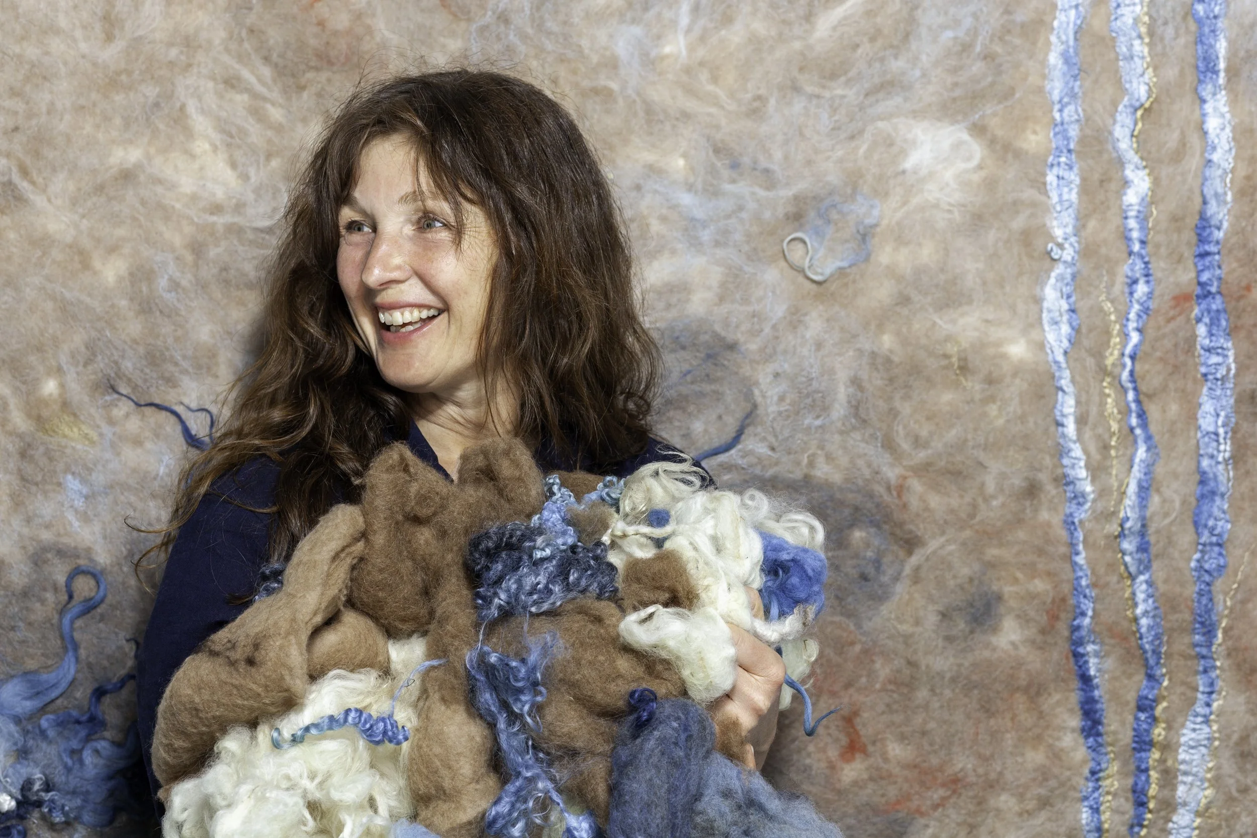 Vrouw met bruin haar die lachend een grote bos wol vasthoudt, omgeven door verschillende gekleurde wol en een achtergrond met textiel en lussen.