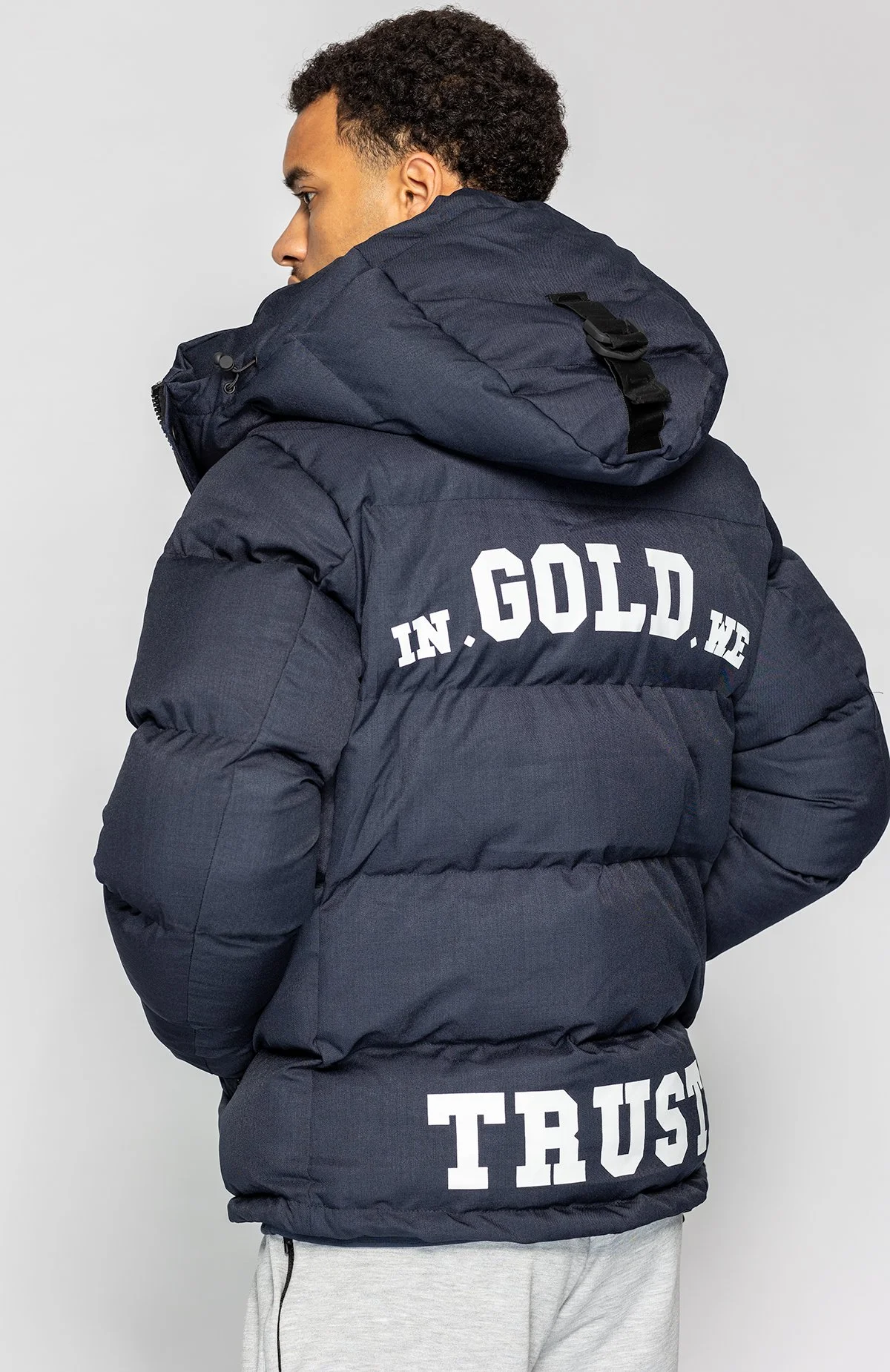 Man met een zwarte gewatteerde jas met witte tekst 'IN GOLD WE TRUST' achterkant