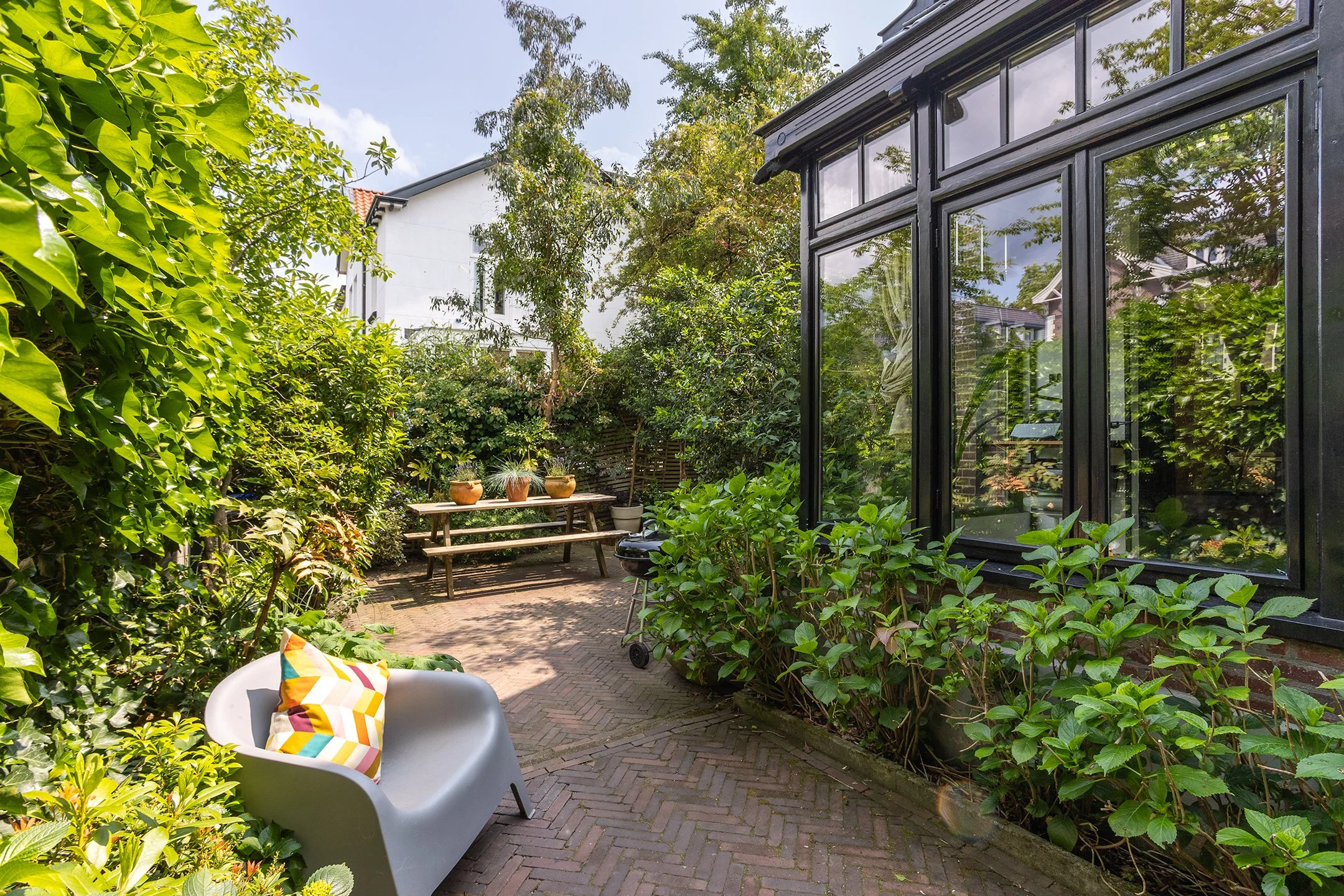 Een gezellige tuin met een bankje met een kleurrijk kussen, omringd door groene planten en struiken, met een glazen kamer van een huis aan de rechterkant en een picknicktafel met potten in de achtergrond.