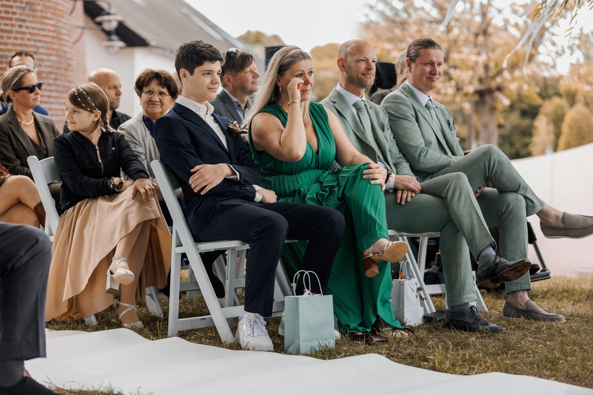 Foto van een groep mensen zittend op witte stoelen buiten tijdens een evenement, met herfstbladeren op de achtergrond.