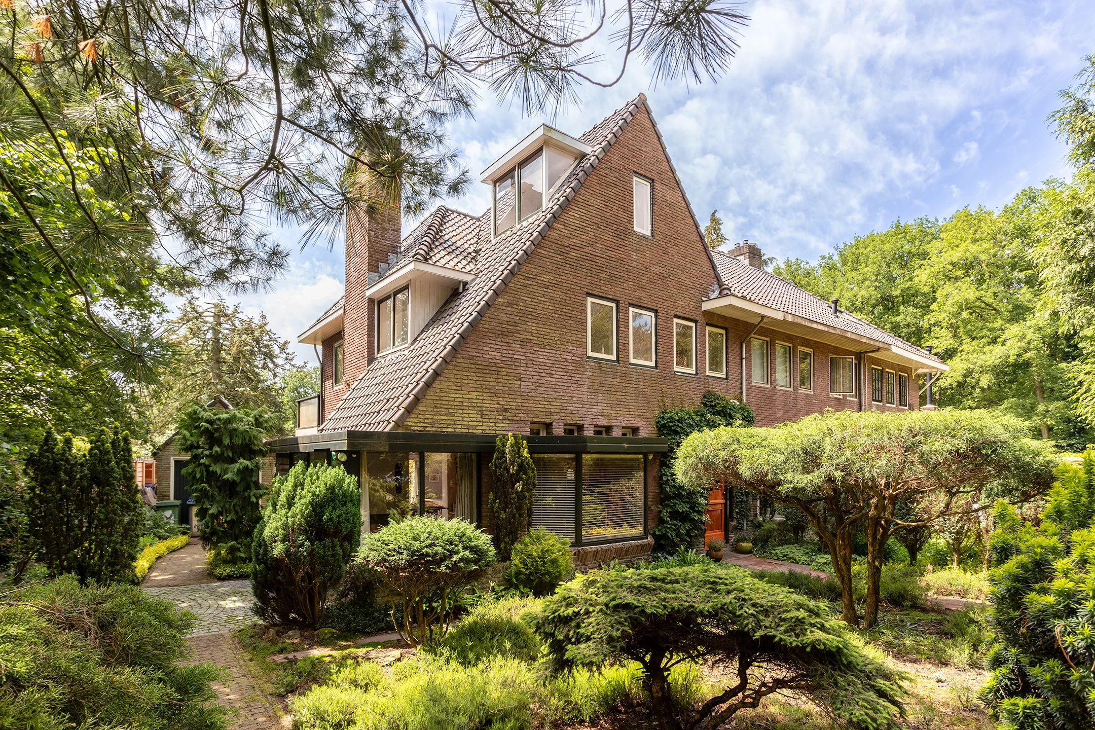 Een huis met een dak van ongelijke hellingen, omgeven door een weelderige tuin met verschillende struiken en bomen, onder een blauwe lucht met wat wolken.