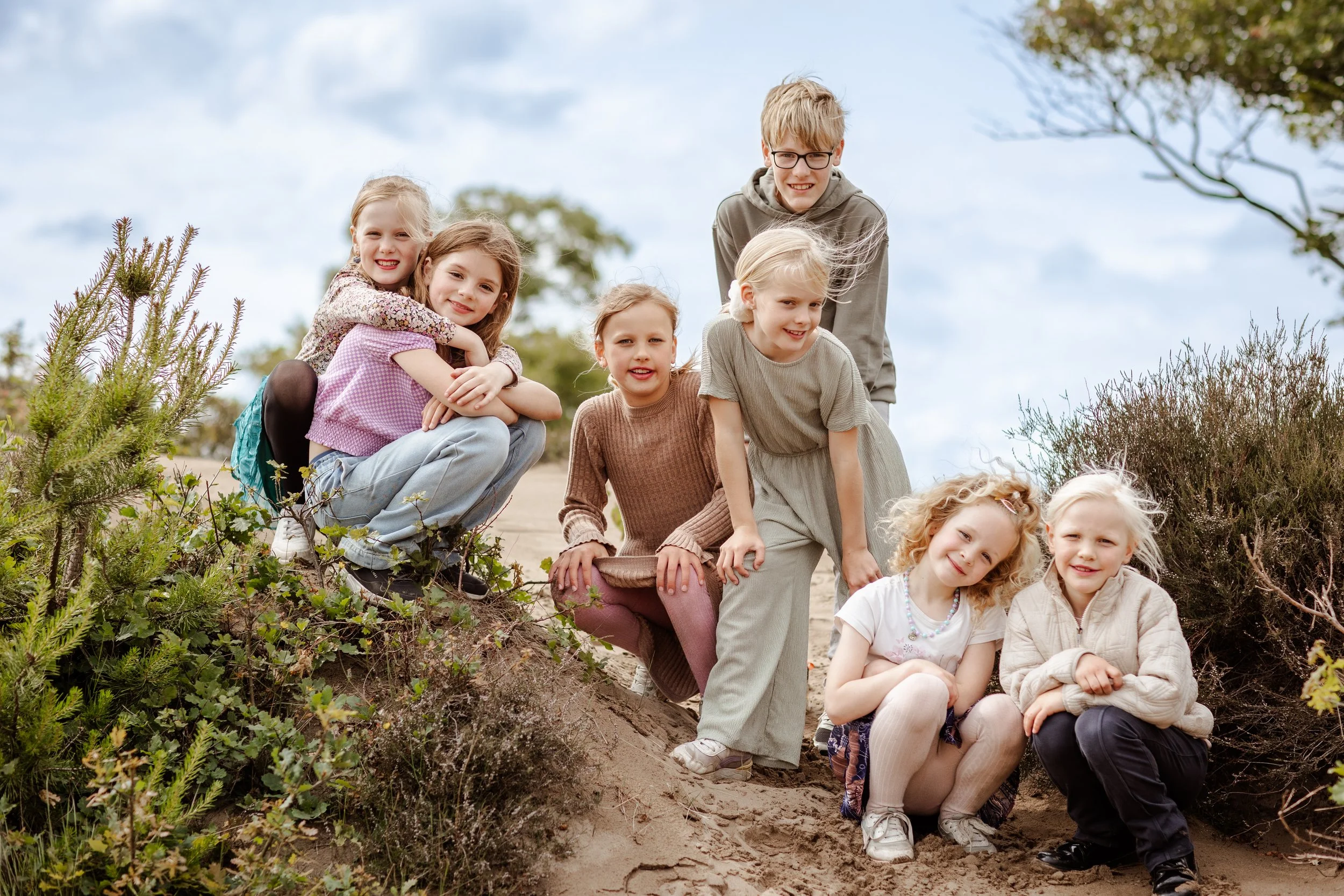 Tien kinderen staan en zitten op een zandheuvel in een buitenomgeving, lachend en poseren voor de camera.