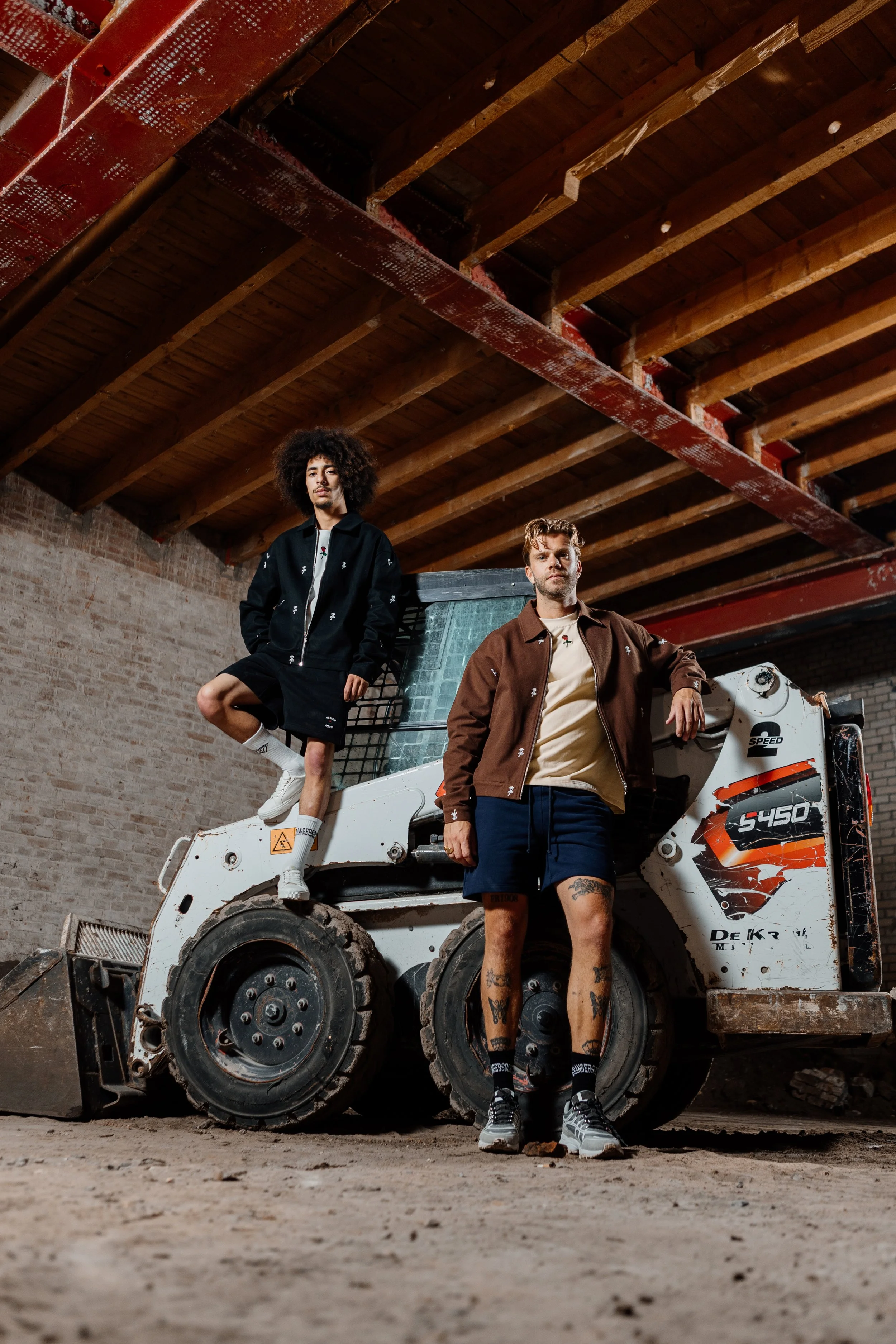 Twee mensen poseren in een constructie-omgeving met een kleine graafmachine, een man staat voorop en een vrouw zit op de machine, beide dragen streetwear en sneakers.