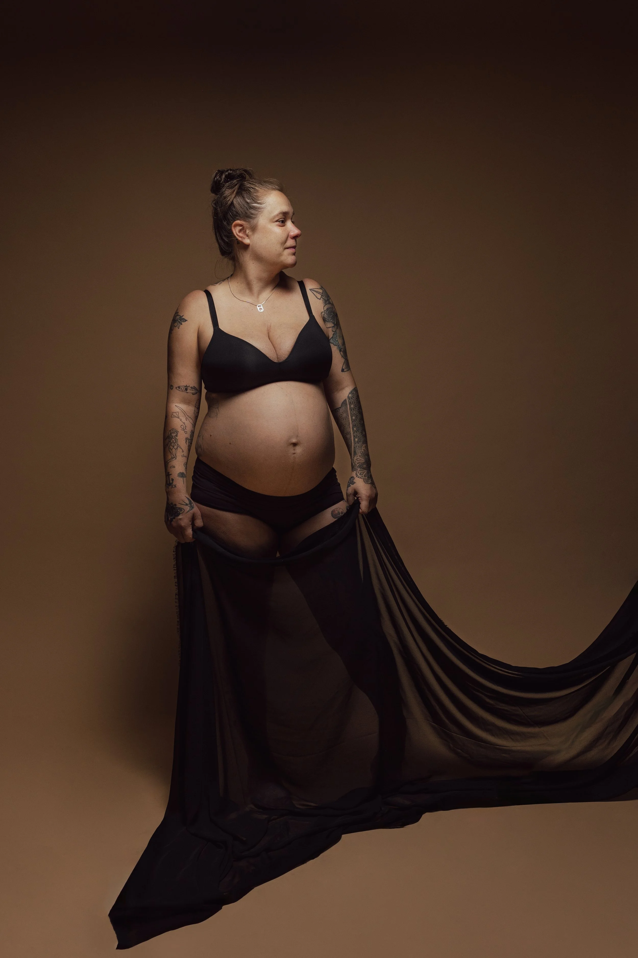 Vrouw met tattoos in zwart lingerie en een lange zwarte stof, zittend tegen een bruine achtergrond.