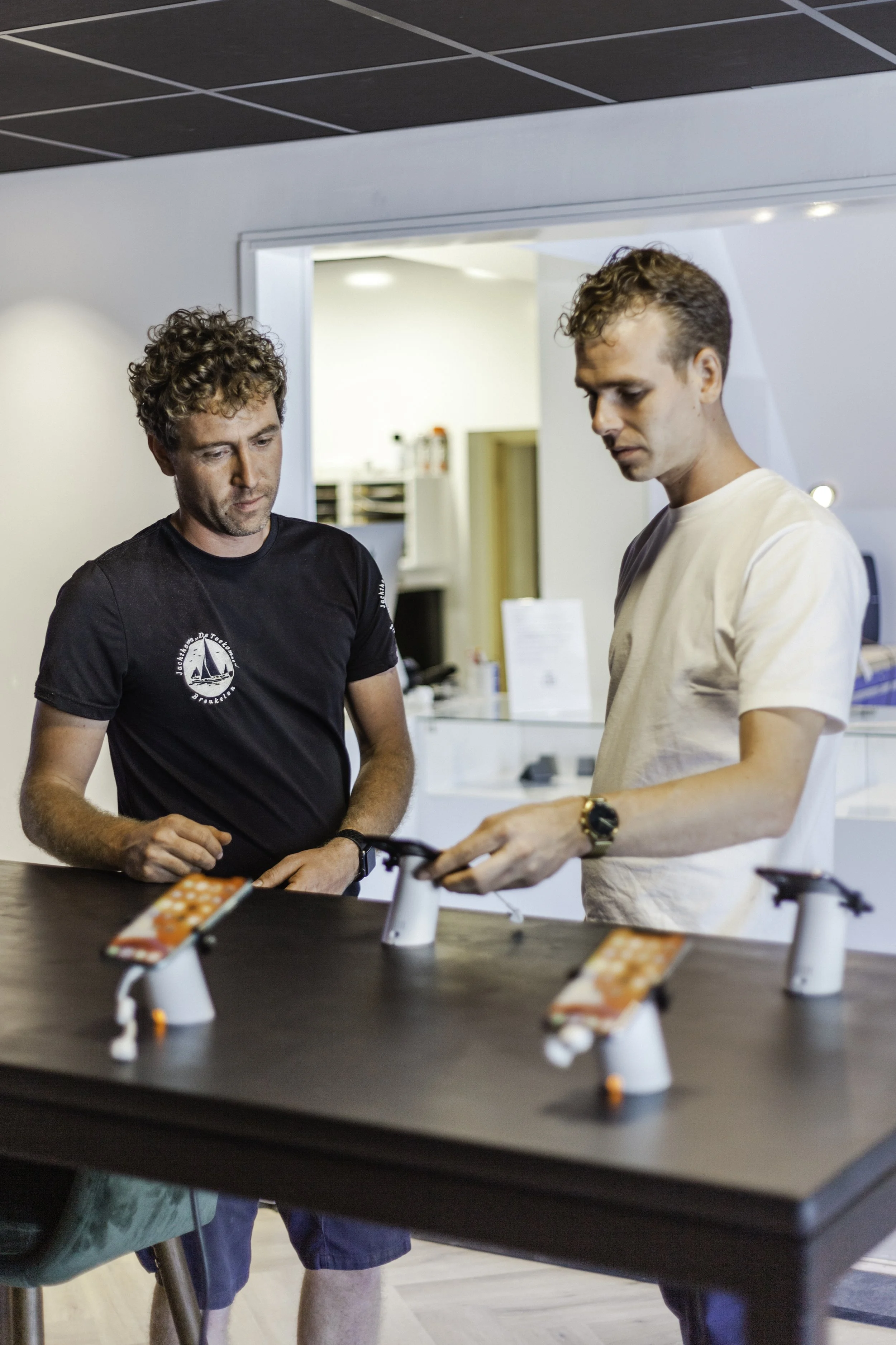 Twee mannen bekijken elektronische apparaten op een tafel in een winkel of showroom.