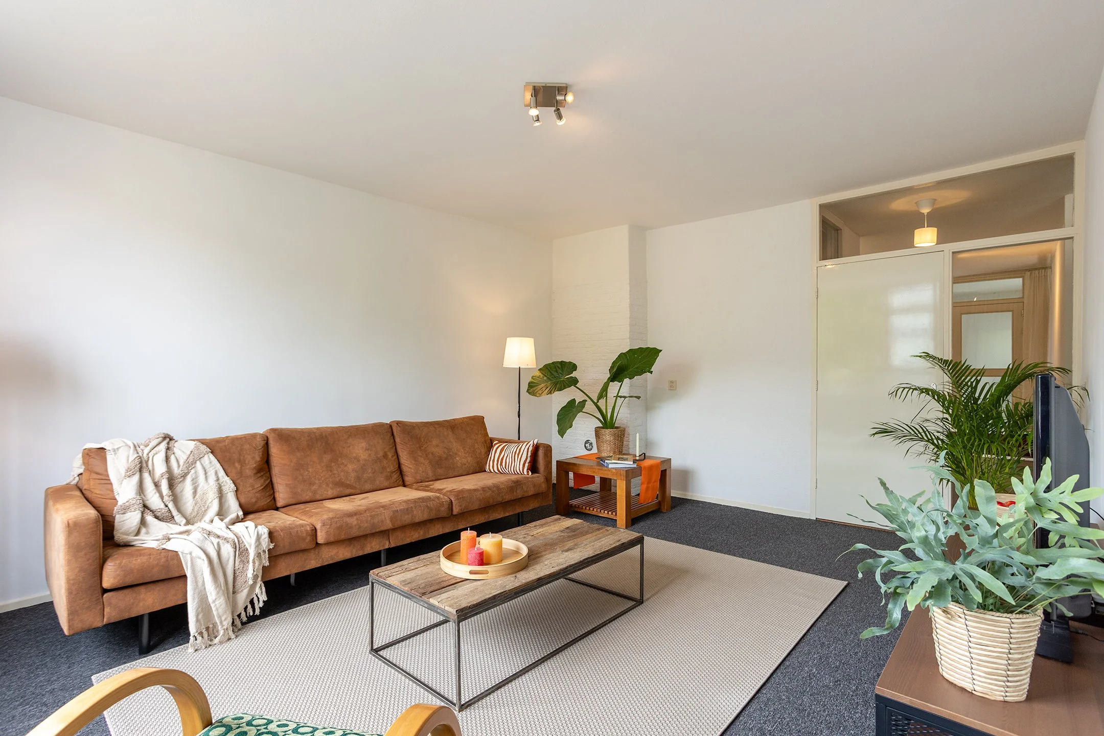 Een woonkamer met een bruin leren bank, een houten salontafel met kaarsen, een vloerlamp en verschillende planten, op een mix van tapijten op een donkere vloer, met witte muren en een tussenmuur met glas