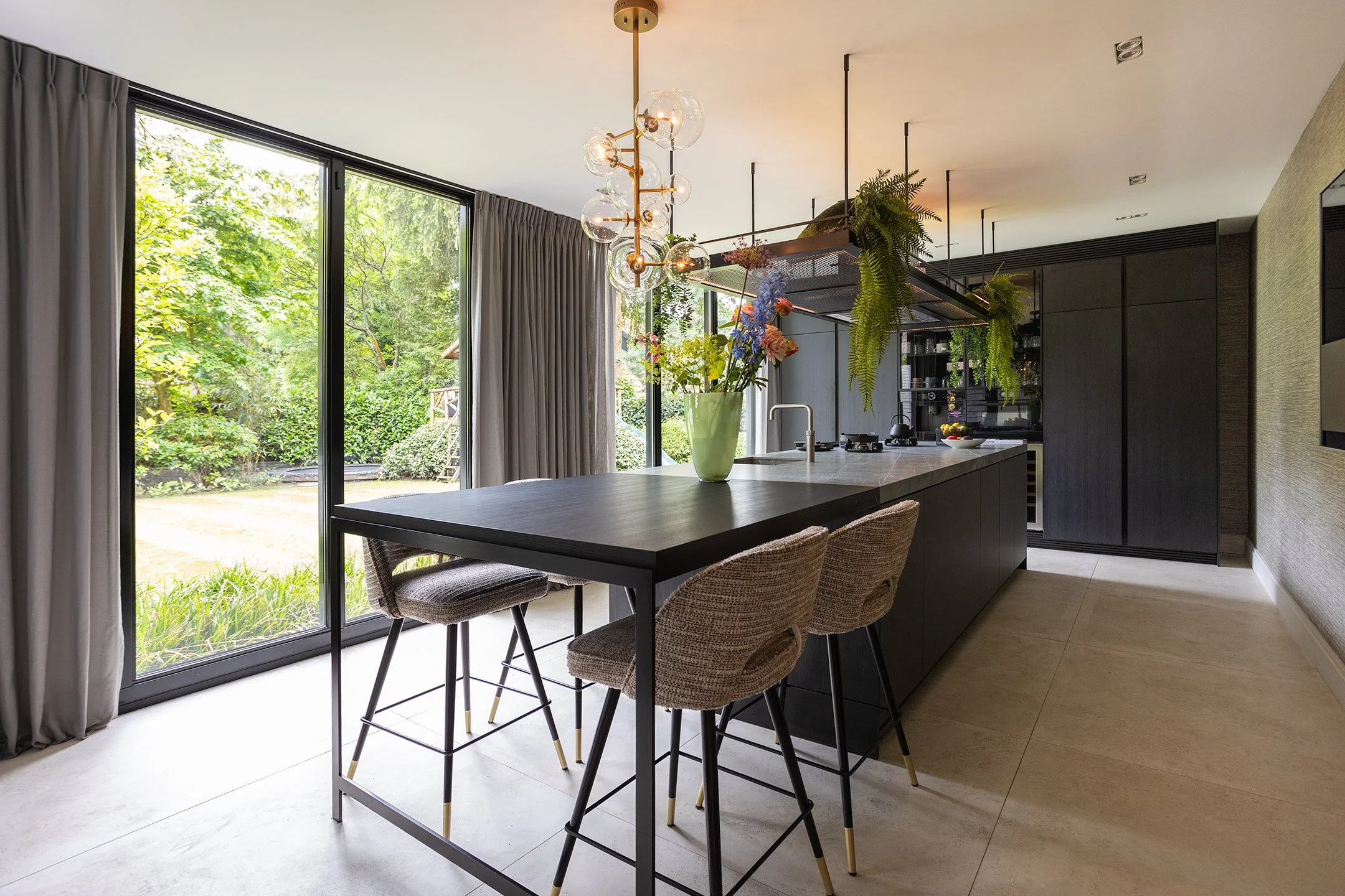 Moderne keuken met zwart keukeneiland, stoelen, glazen schaal met bloemen, grote ramen met grijze gordijnen en groene tuin buiten.