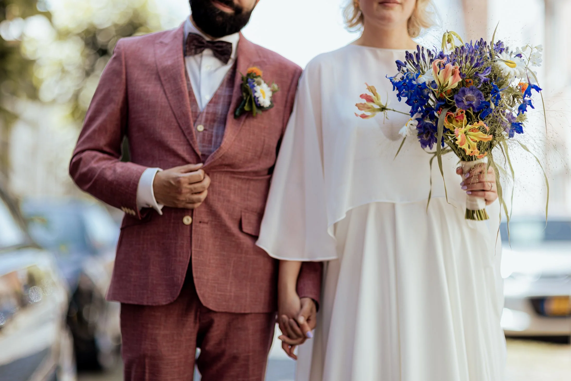 Een getrouwd paar houdt hand in hand, de vrouw houdt een kleurrijke bos bloemen, de man draagt een roodbruin pak met een bloemenboutonnière.