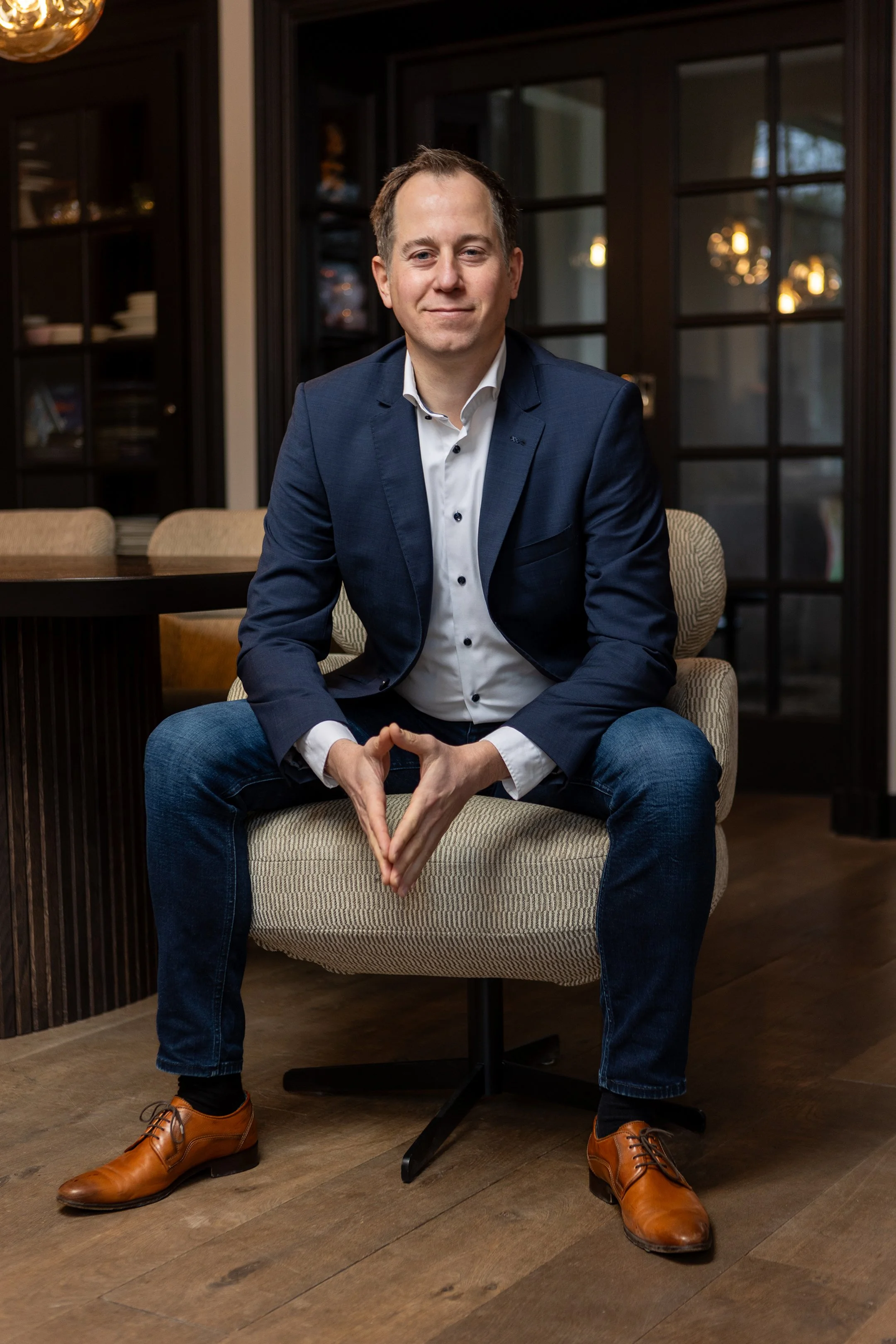 Man in een blauwe blazer en witte blouse zittend op een kantoorstoel in een kantoor met houten vloer en glazen deuren op achtergrond.