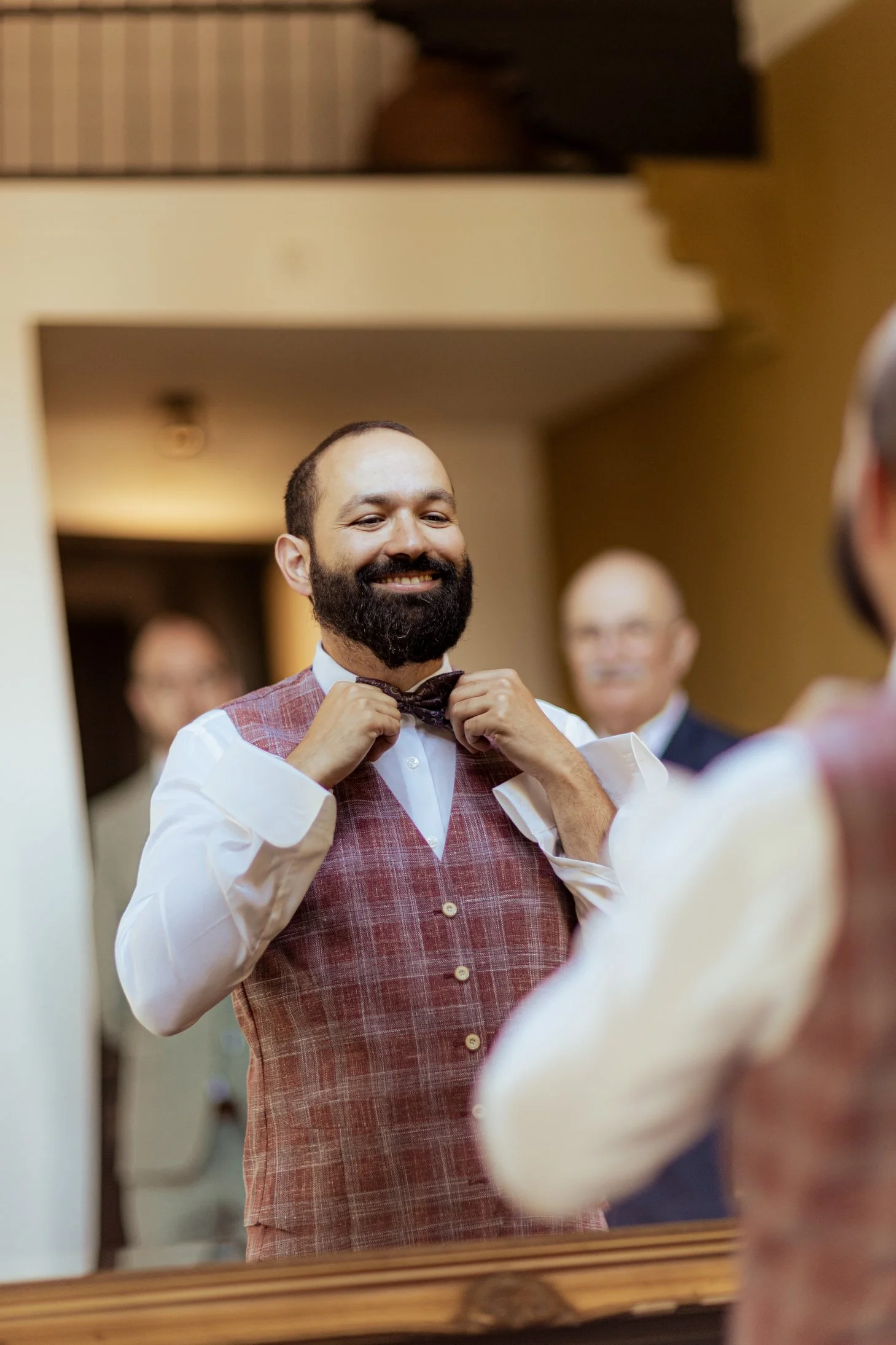 Man die zichzelf in de spiegel mooi maakt, met een glimlach, terwijl hij zijn stropdas rechtzet, in een huis met vrienden of familie op de achtergrond.