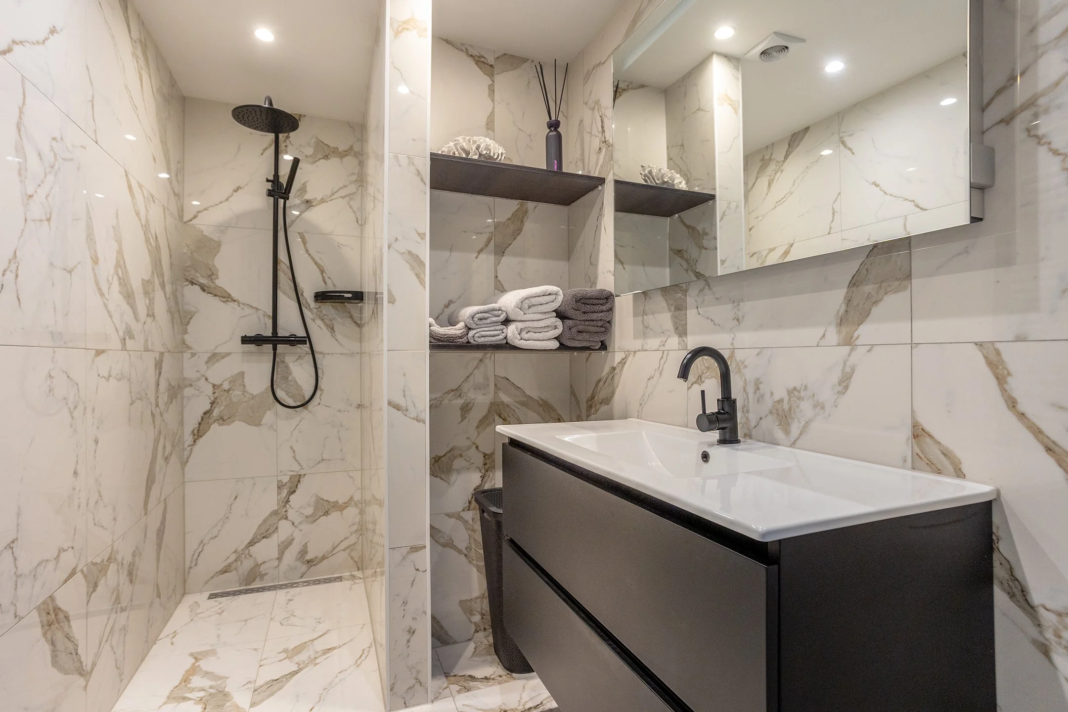Een moderne badkamer met marmeren wand- en vloertegels, een inloopdouche met een zwarte regendouche en handdouche, een zwart wastafelmeubel met een witte wastafel, spiegels en open schappen met handdoeken en decoratie.