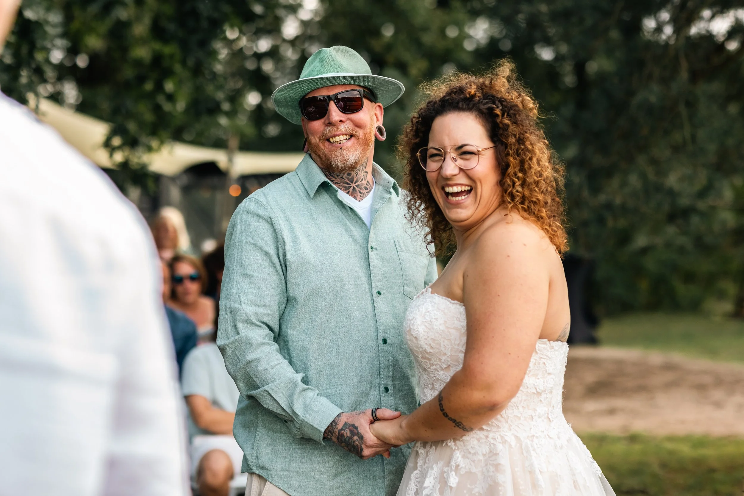 Treurend bruid en bruidegom die elkaar vasthouden tijdens hun huwelijk buiten, lachend en plezier makend, met gasten op de achtergrond.
