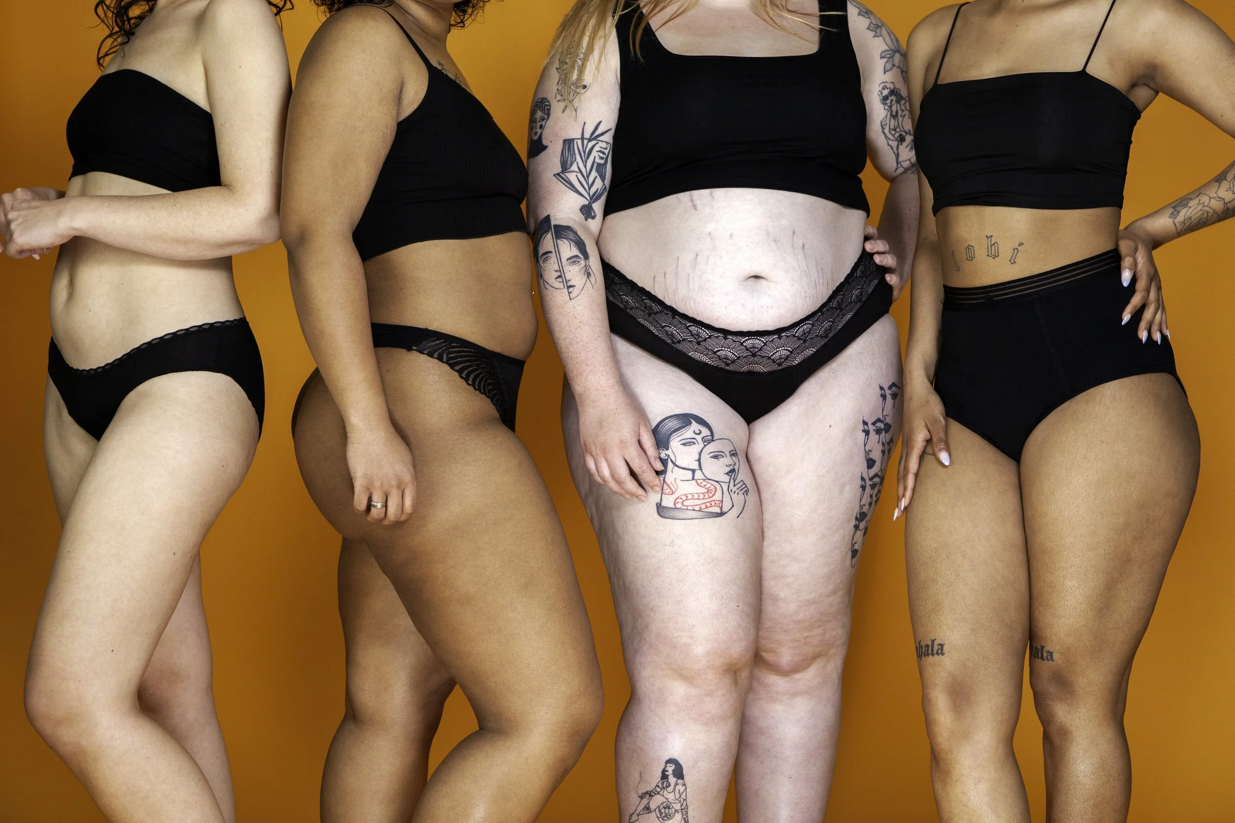 Vijf vrouwen in zwarte onderkleding staan tegen een oranje achtergrond. Ze tonen hun tattoo's en lichaamsvormen.