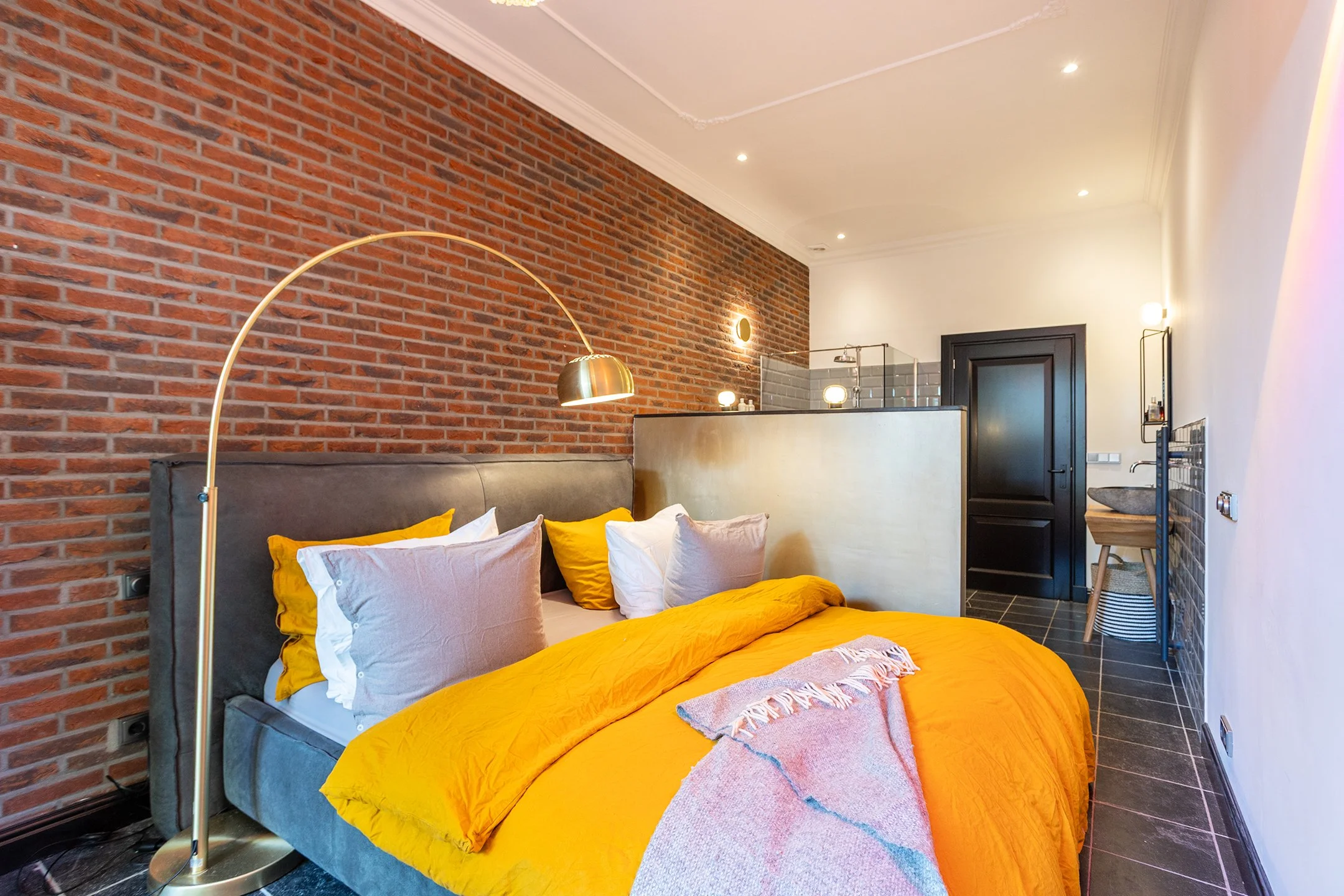 Een slaapkamer met een bed dat een gele dekbedovertrek en meerdere kussens heeft, tegen een bakstenen muur, met een goudkleurige booglamp erboven.