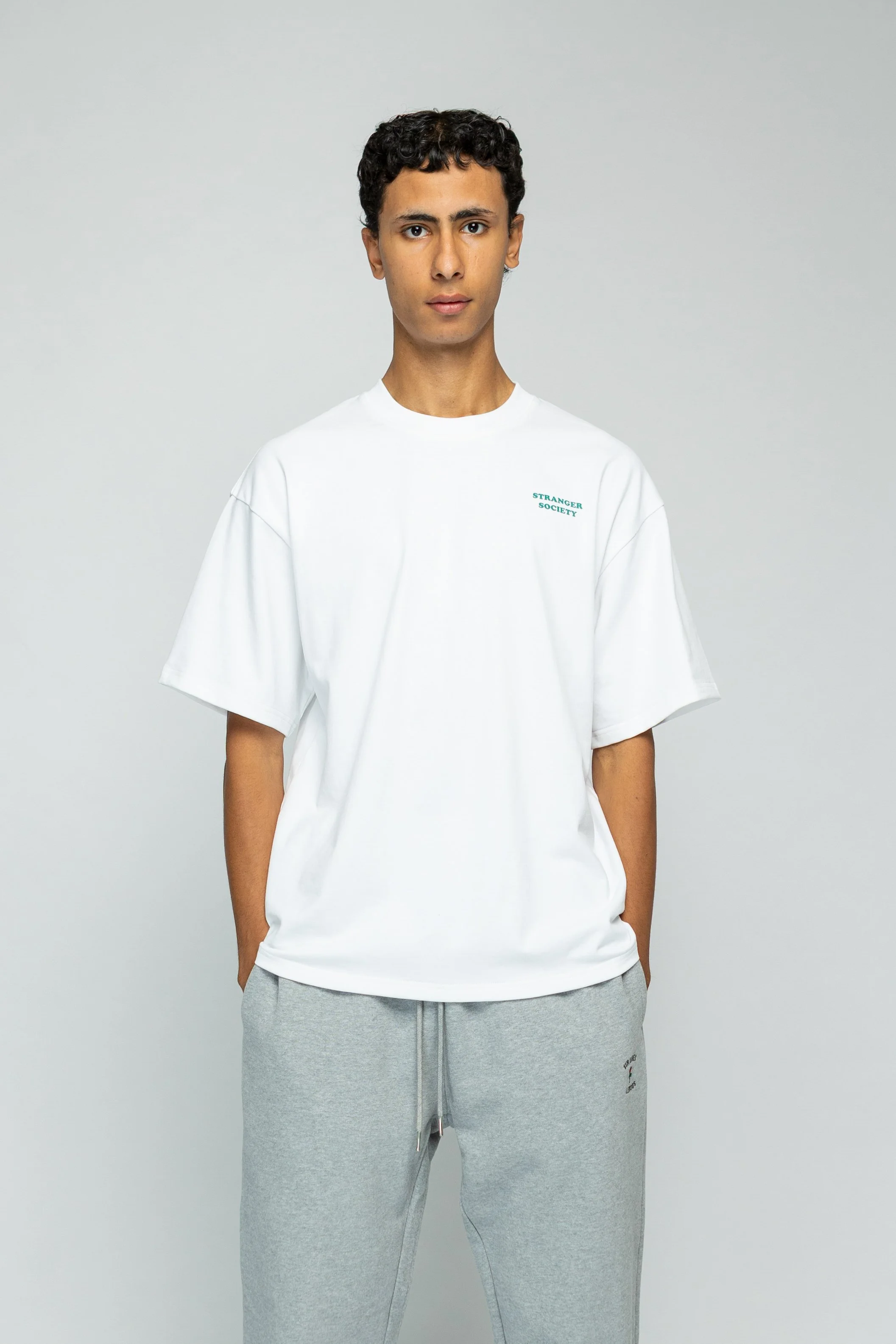 Man staat rechtop tegen een effen witte achtergrond, draagt een witte T-shirt met de tekst 'Stranger Society' en grijze joggingbroek.