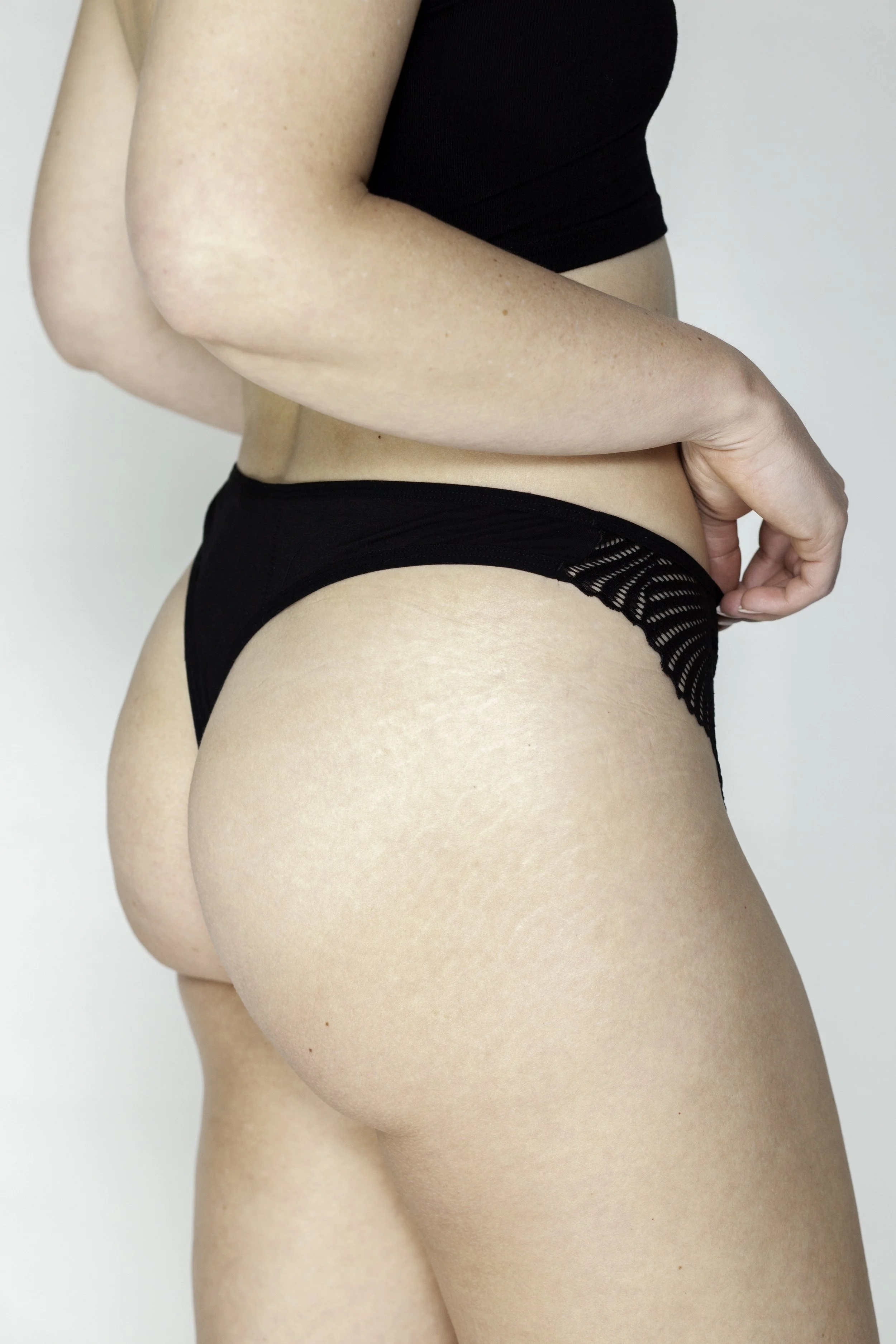 Close-up van een vrouw in zwart ondergoed, haar hand rust op haar heup, tegen een lichte achtergrond.