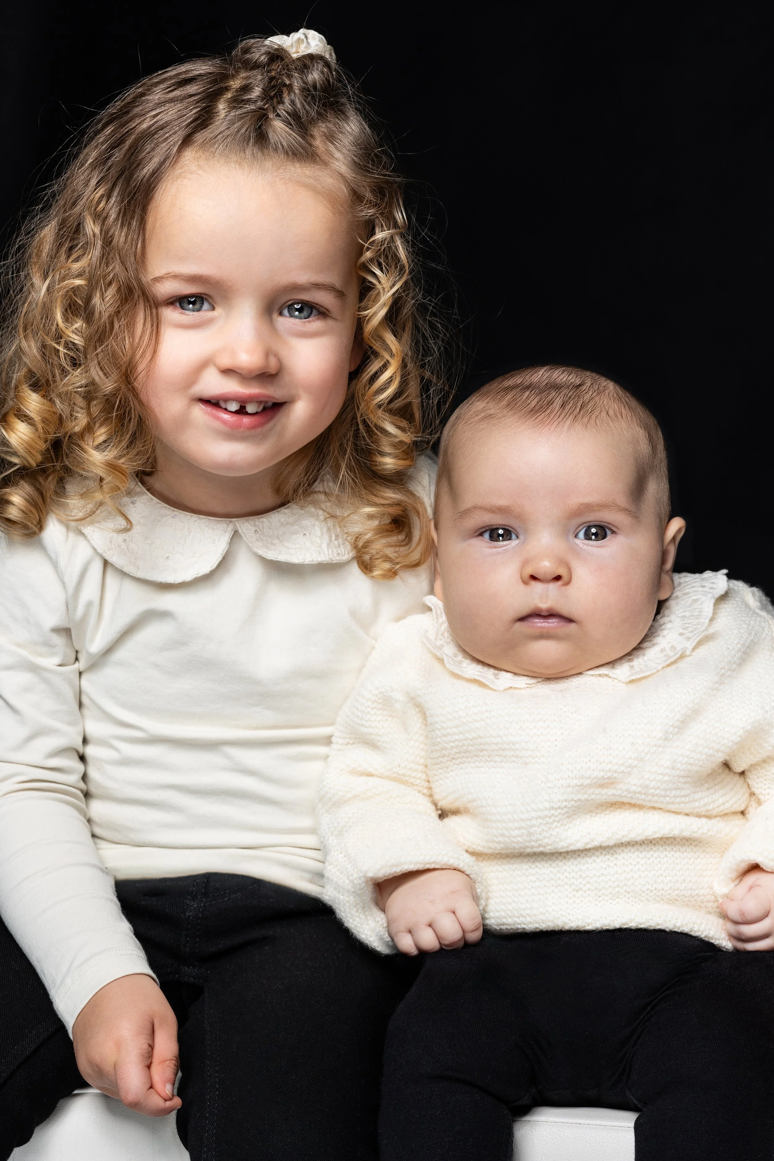 Twee jonge kinderen, een meisje met krullend haar en een glimlach, en een baby, zitten samen tegen een zwarte achtergrond.