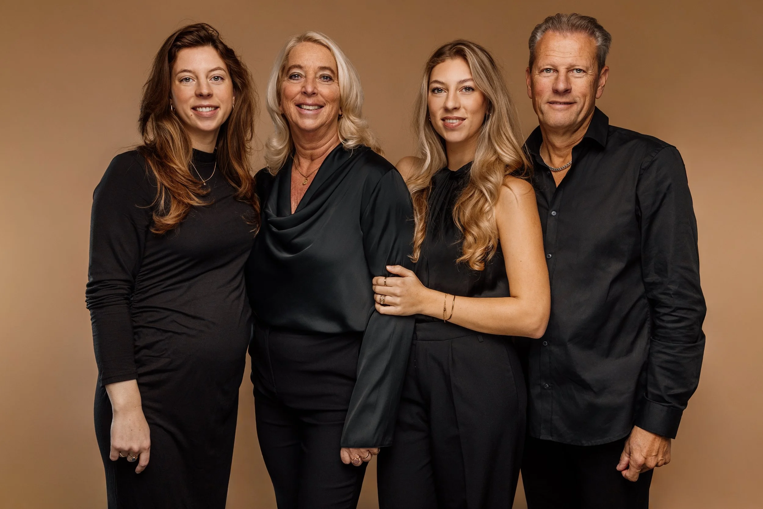 Vier mensen, drie vrouwen en een man, staan zij aan zij tegen een beige achtergrond. Ze dragen allemaal zwarte kleding, lachen en kijken naar de camera.