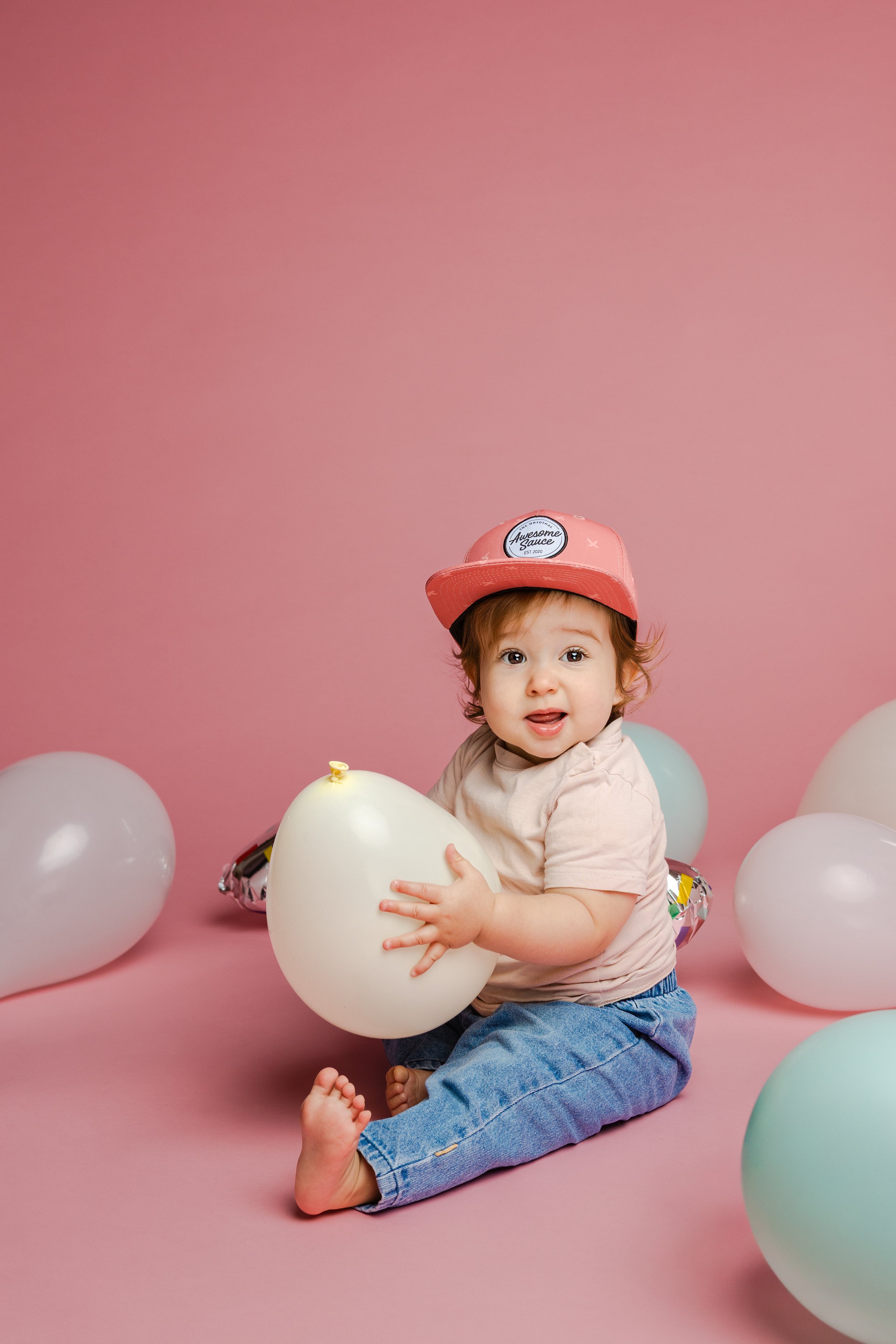 Een schattige baby met een roze pet die op een roze achtergrond zit, omgeven door witte en pastelgekleurde ballonnen, terwijl hij een witte ballon vasthoudt.