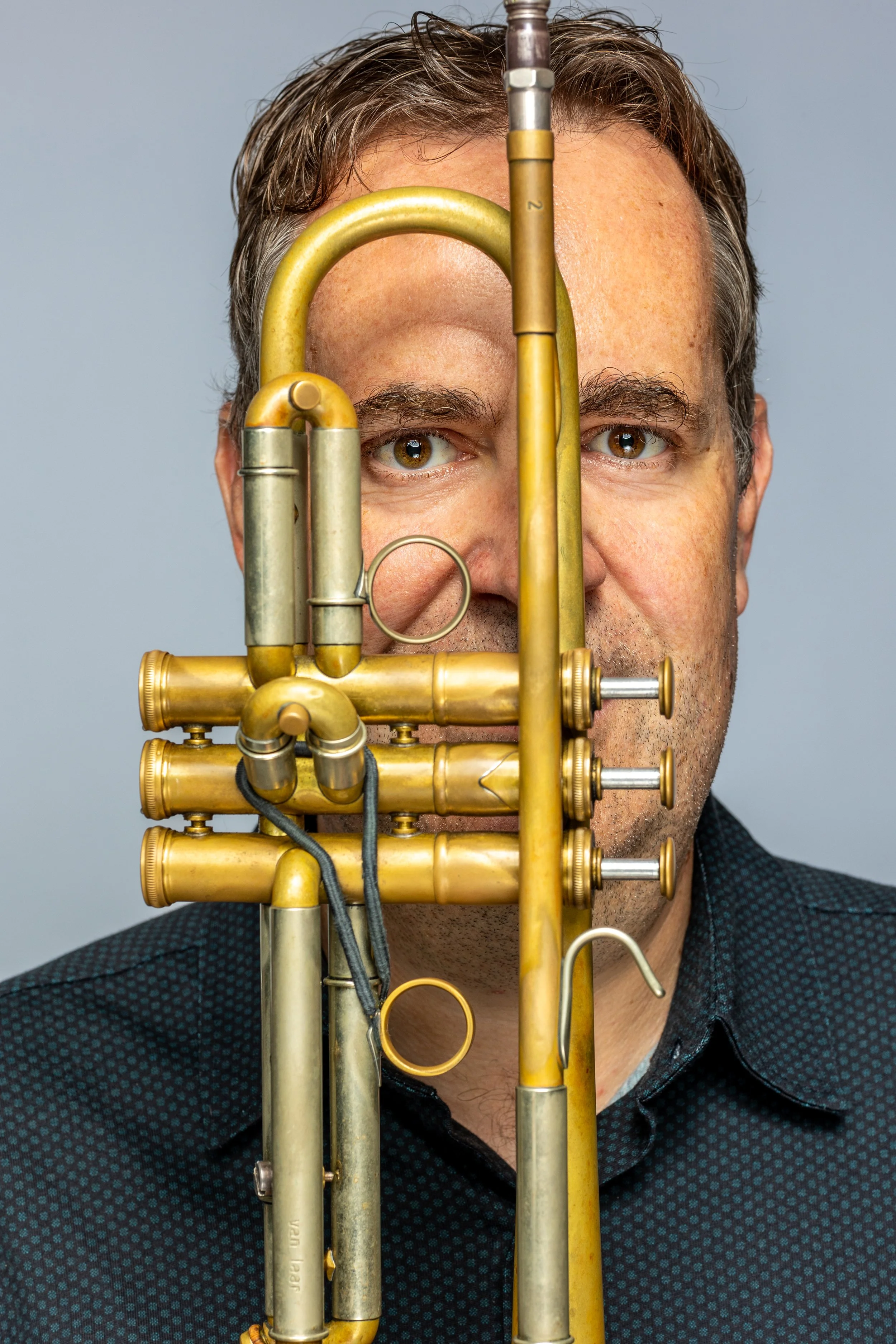 Een man met een snor en korte haar, die achter een oude, gouden trompet staat. De trompet bedekt ongeveer de helft van zijn gezicht, waardoor alleen zijn ogen en een deel van zijn mond zichtbaar zijn.