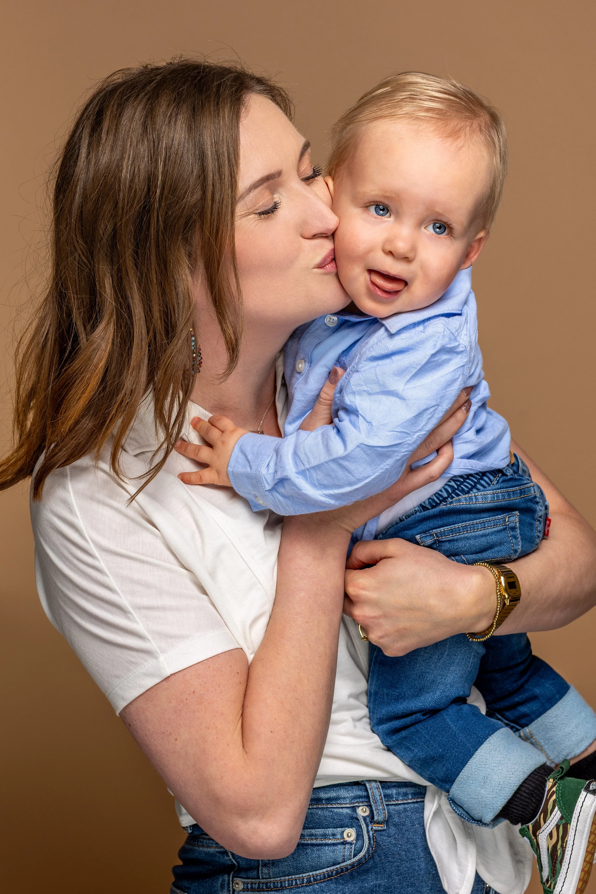 Vrouw kust een lachende peuter op de wang terwijl hij met zijn tong uitsteekt, tegen een effen bruine achtergrond.