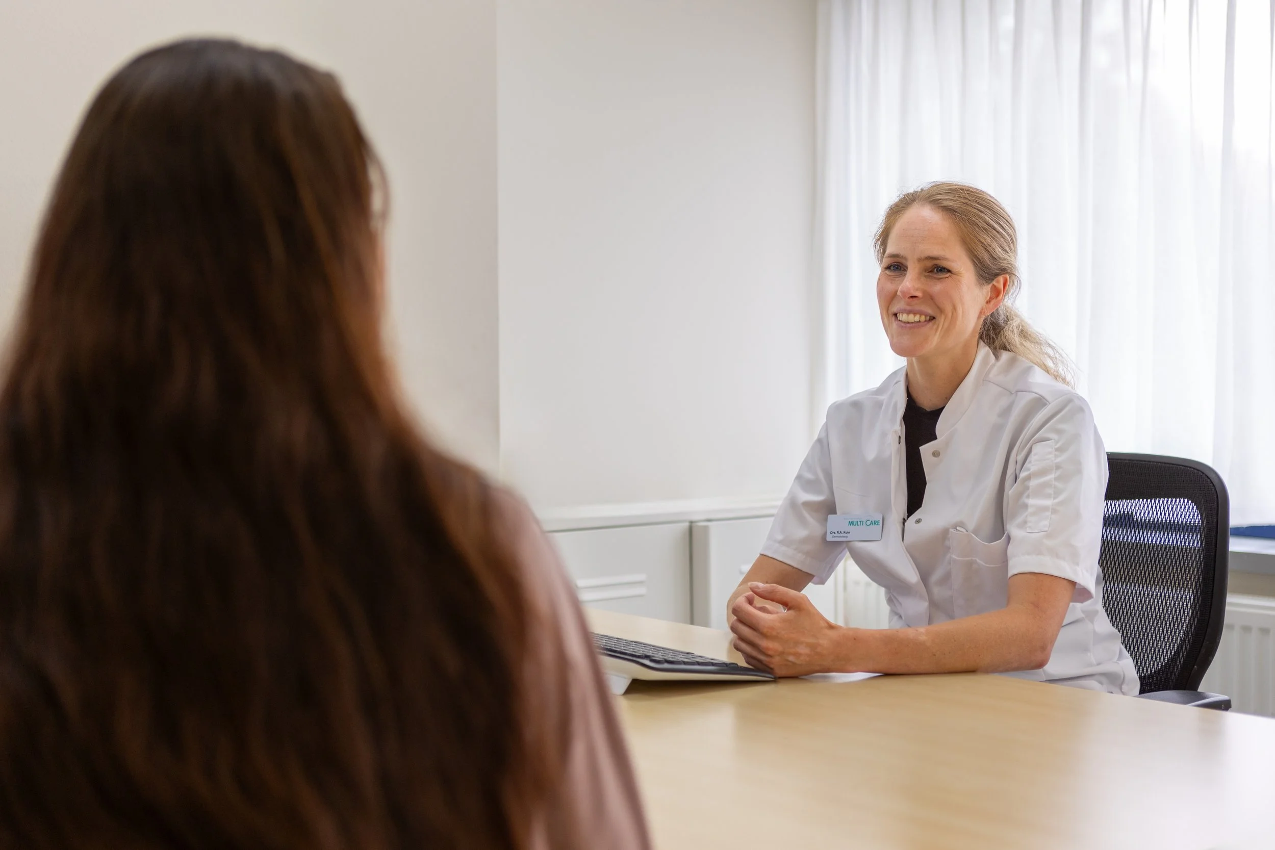 Een vrouw in een witte doktersjas zit aan een bureau en praat met een patiënt, die zichtbare bruine haar heeft. De dokter glimlacht en heeft haar handen gevouwen op het bureau.