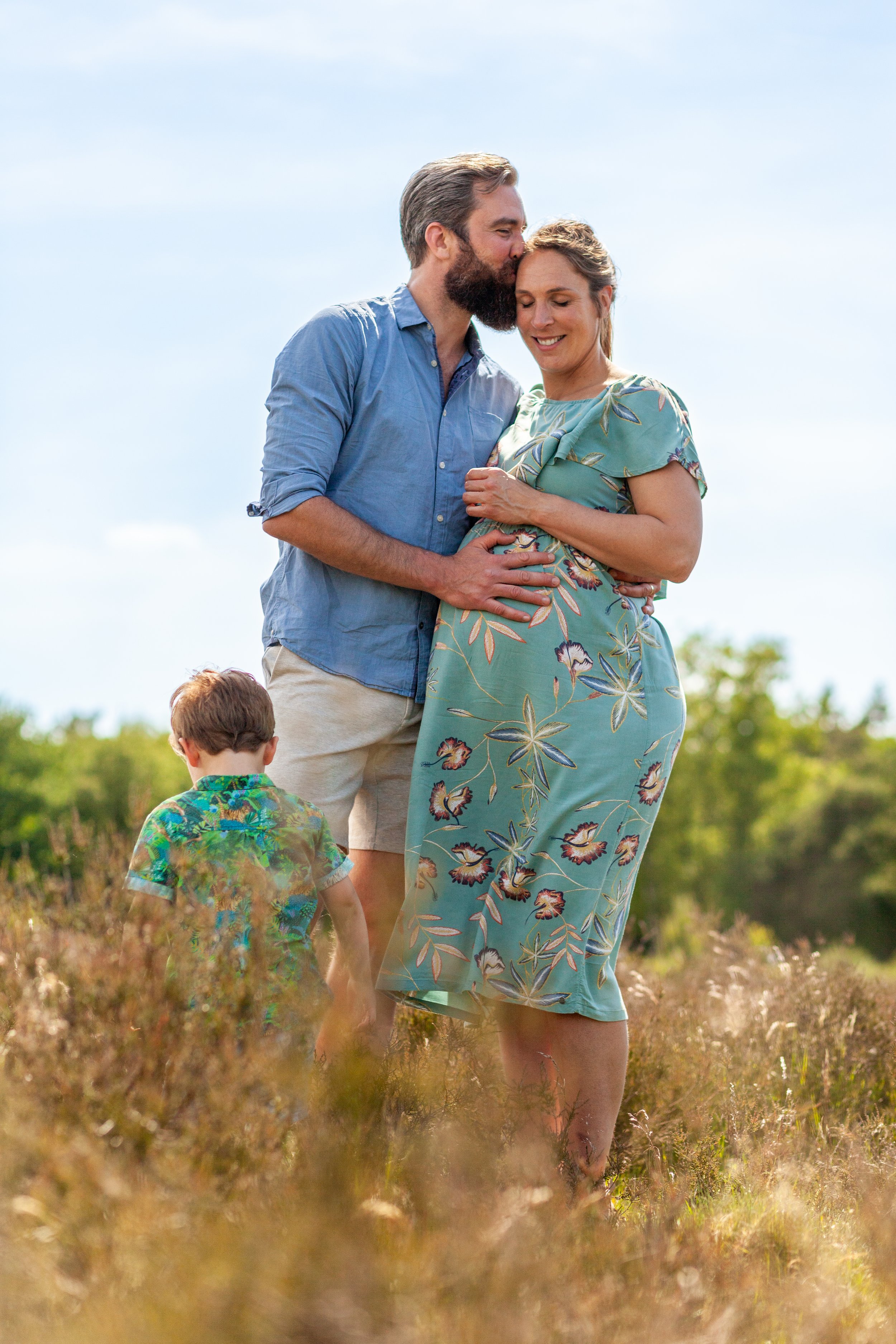 Een zwangere vrouw en haar partner geven elkaar een kus op het voorhoofd in een veld, met een kind dat wegloopt op de voorgrond.