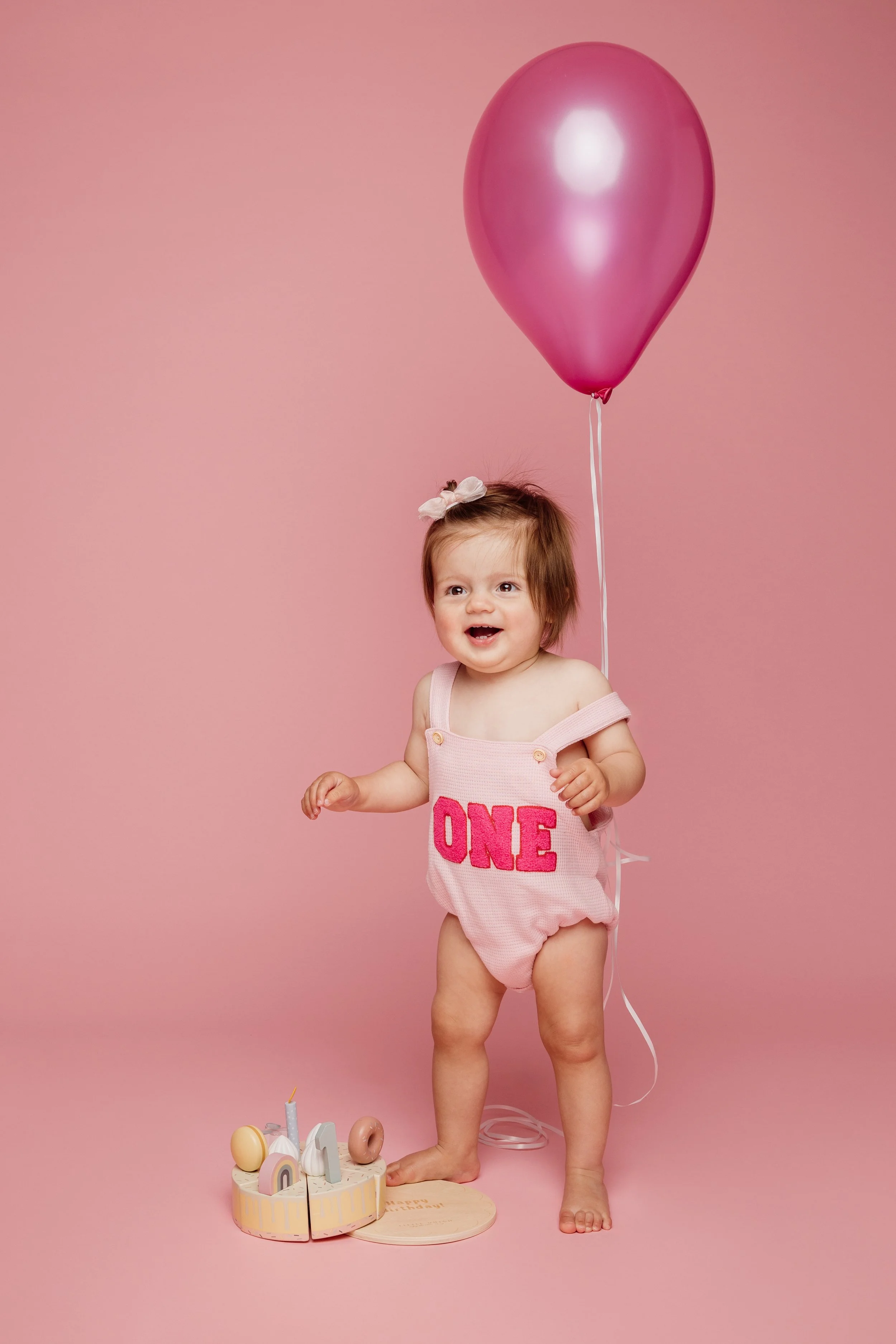 Een jong meisje met een bloesem haarclip, gekleed in een roze romper met de tekst 'ONE', staat naast een verjaardagstaart en houdt een roze ballon vast tegen een roze achtergrond.