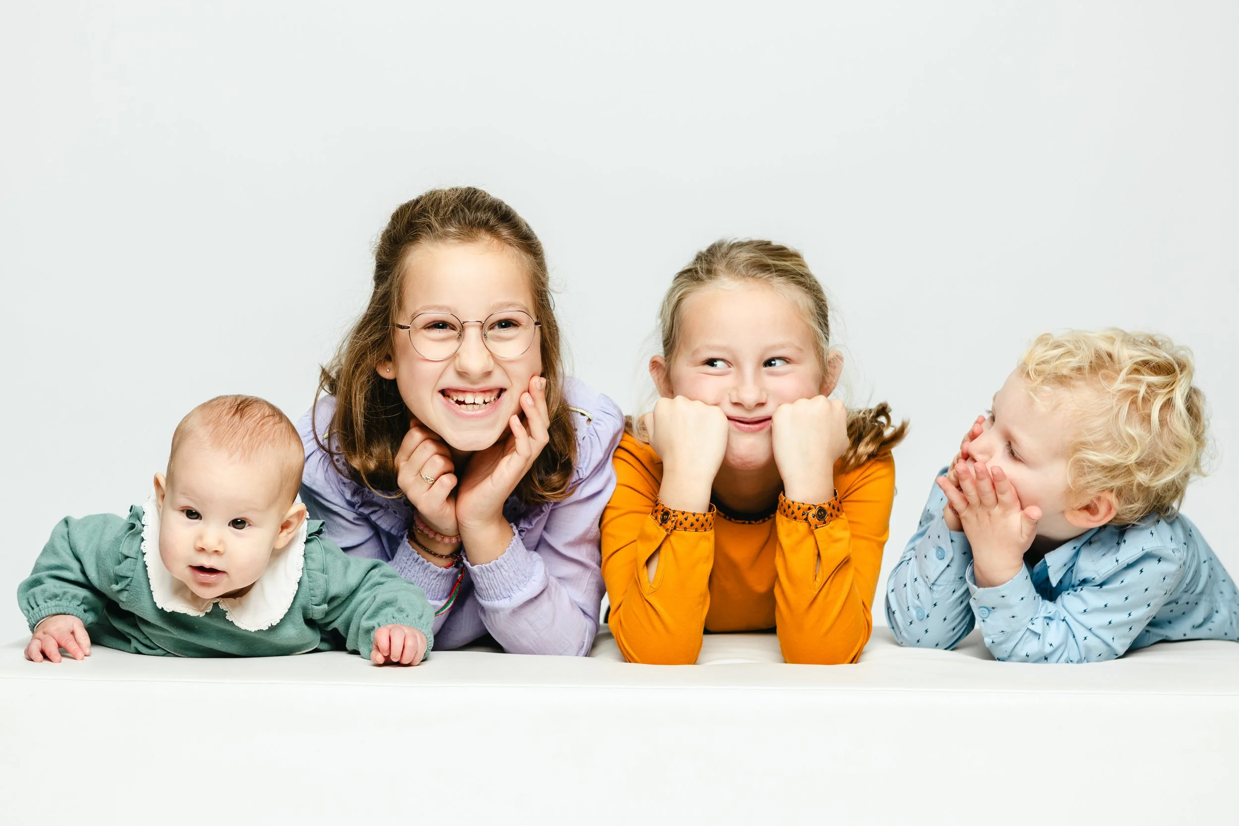 Vier kinderen liggen op hun buik tegen een witte achtergrond, lachen en maken grappen. 