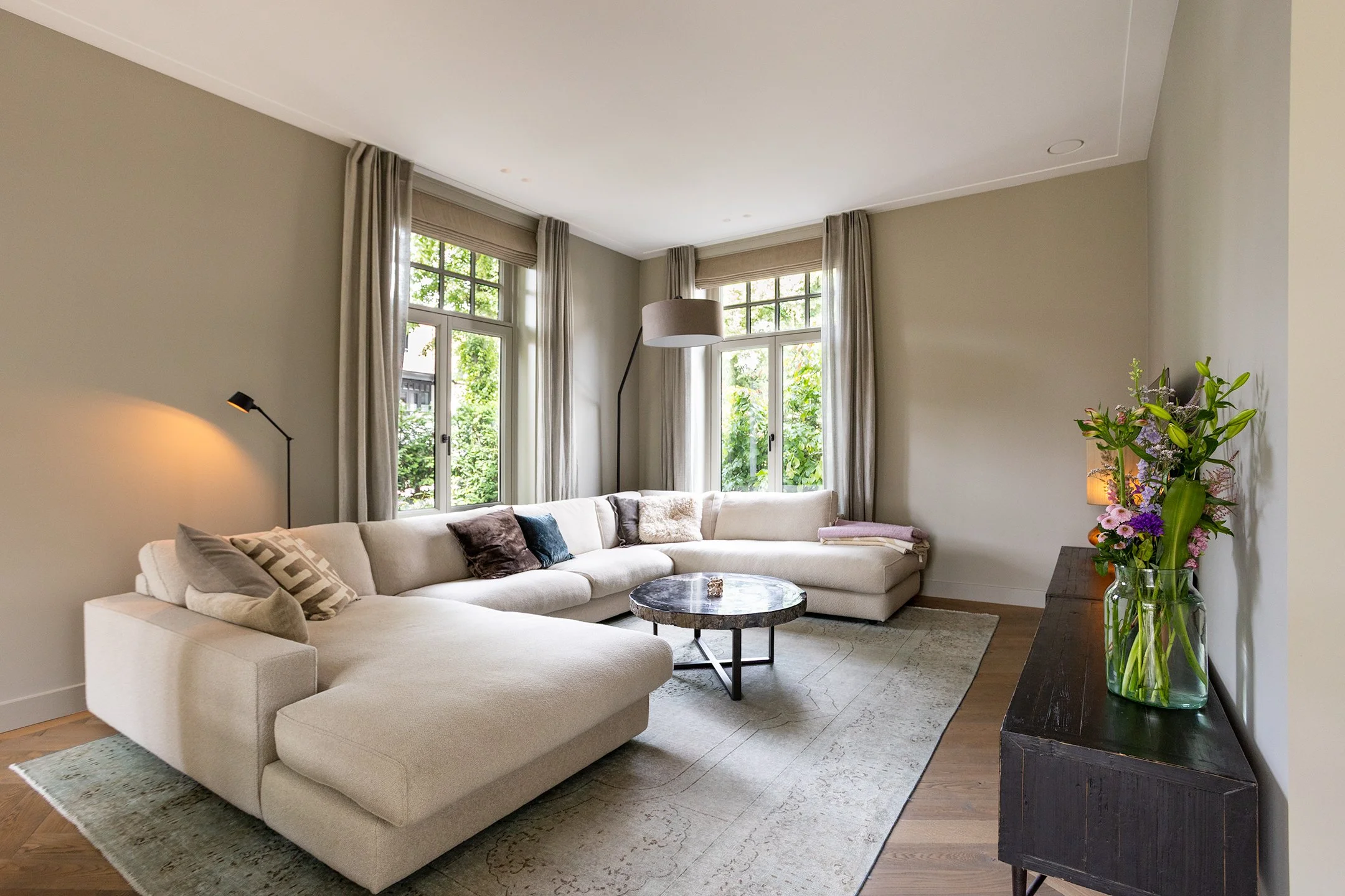 Lichte woonkamer met een grote L-vormige witte bank, een ronde marmeren salontafel, een zwarte houten kast met een vaas met bloemen, grote ramen met beige gordijnen, een vloerlamp en een plafondlamp.