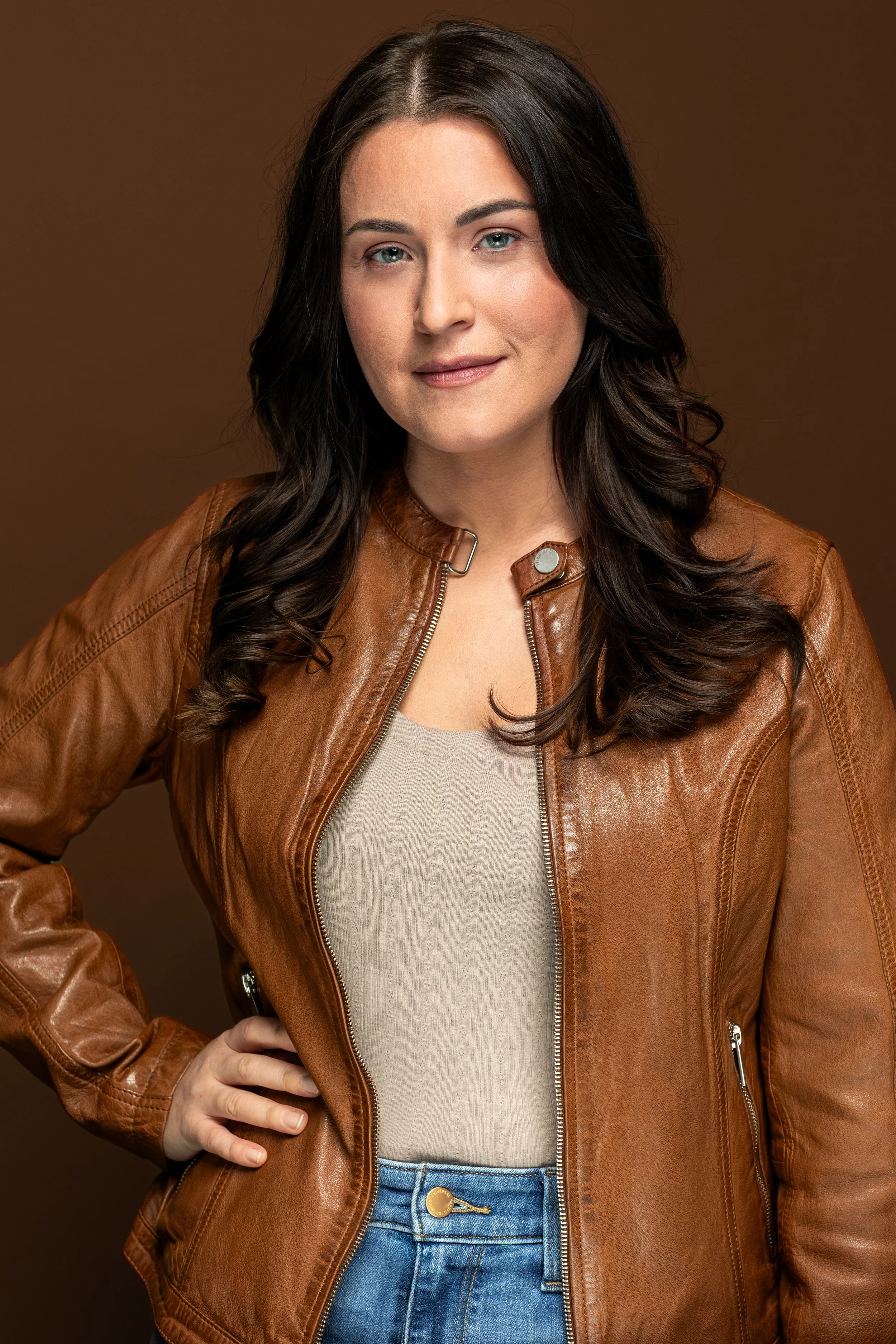 portret van een vrouw met lang, donker haar, blauwe ogen, een lichte huid, die lacht en een bruine leren jas draagt, staand tegen een bruine achtergrond