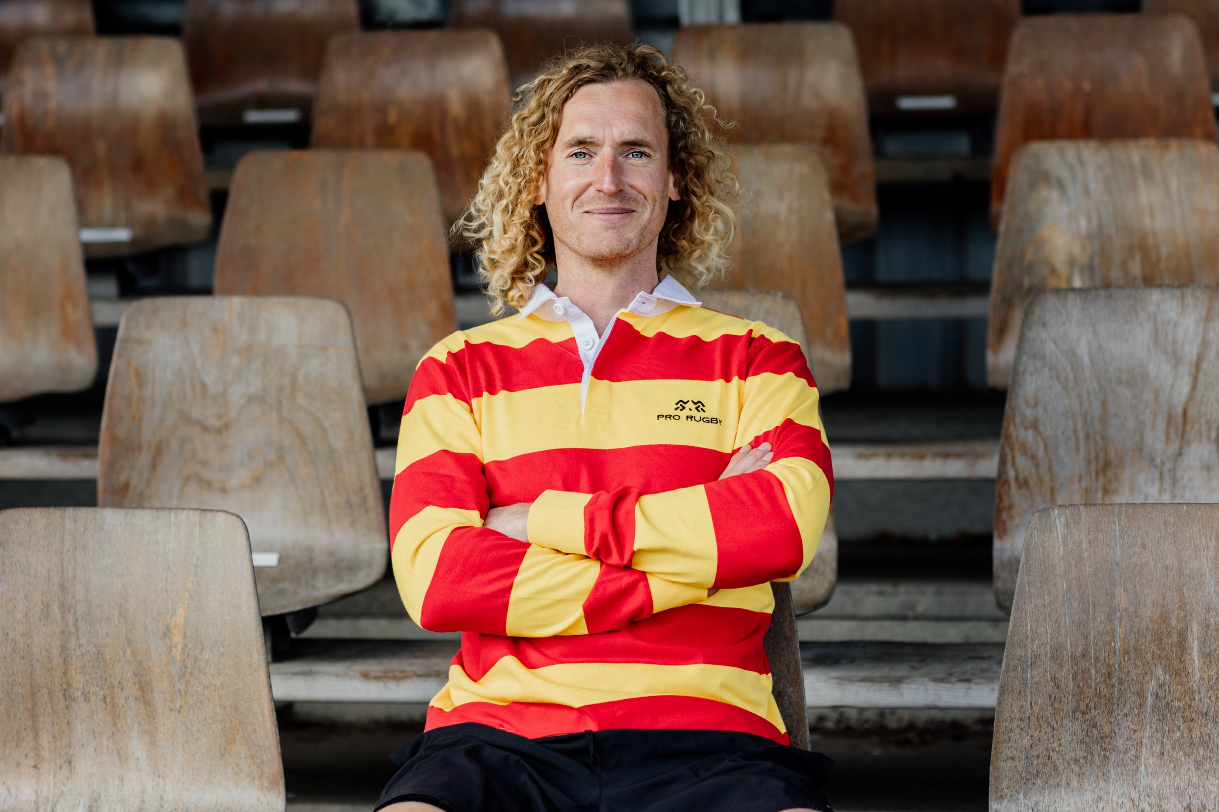 Man met lang, gekruld blond haar zit op een stoel op een rugbytribune, gekleed in een rood en geel rugbyshirt met een witte kraag.
