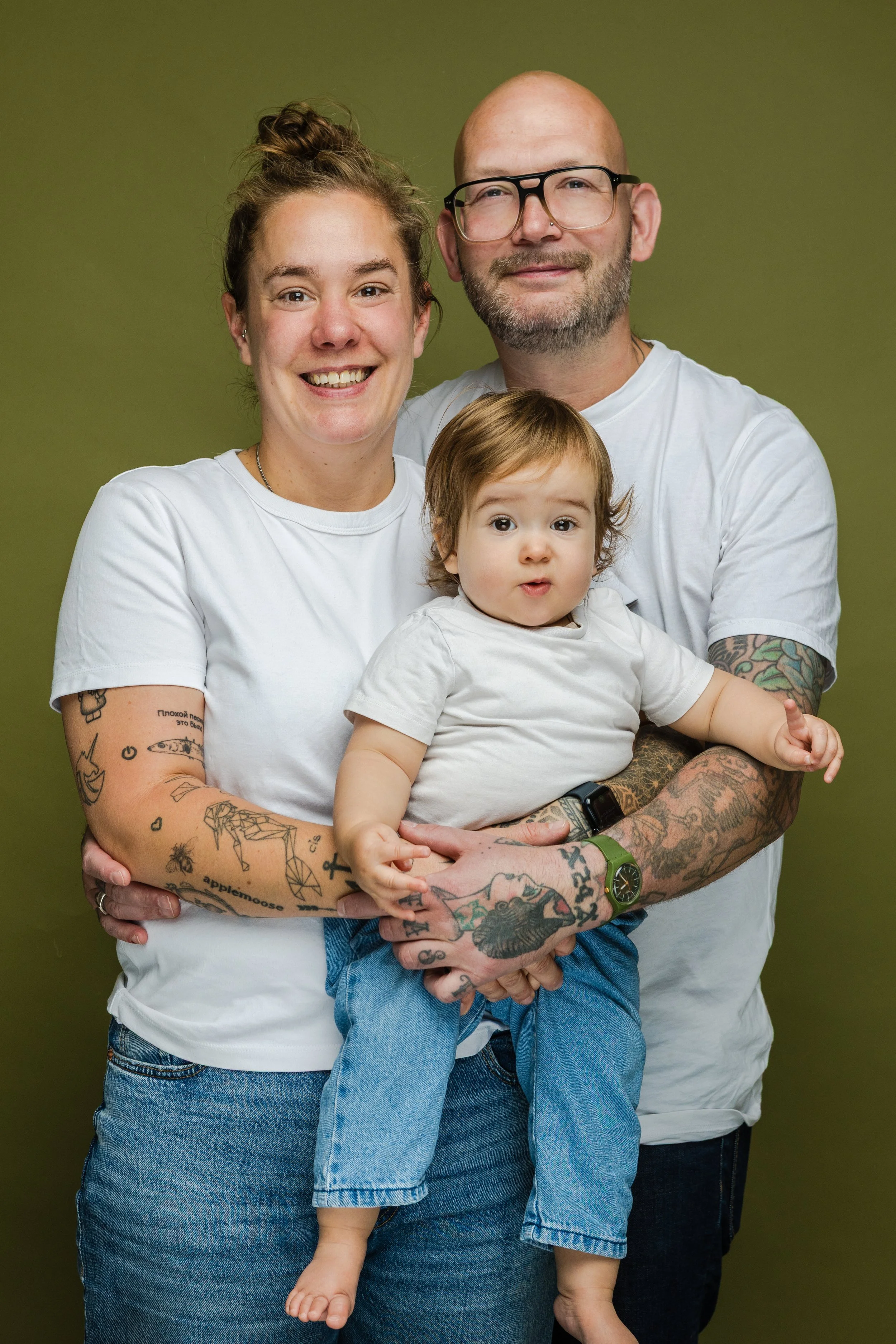 Gezinsfoto met een vrouw, een man en een klein kind, allemaal lachend voor een groene achtergrond.