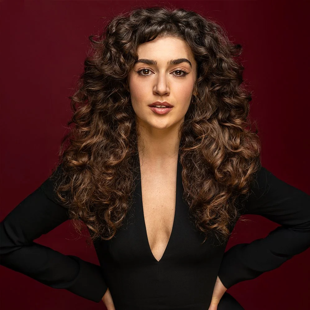 Portret van een vrouw met lange, krullende bruin haar, gekleed in een zwarte jurk met een diepe V-uitsnijding, poseert voor een donkere rode achtergrond.