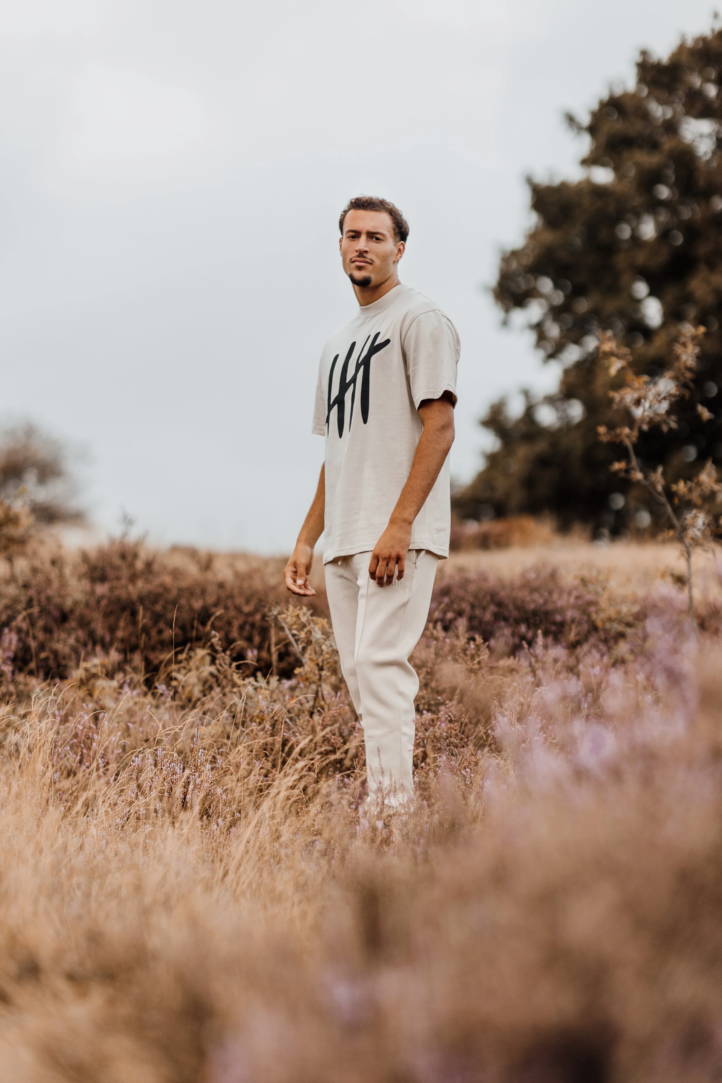 Man staat in een veld met droog, bruin gras en bloemen, draagt een beige T-shirt met zwarte tekens en witte broeken, onder een bewolkte hemel.