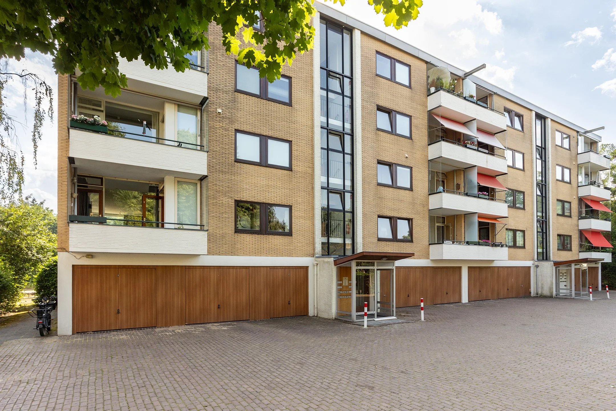 Een modern appartementengebouw met meerdere verdiepingen, grote ramen en balkons, omgeven door groen en een parkeerplaats aan de voorkant.