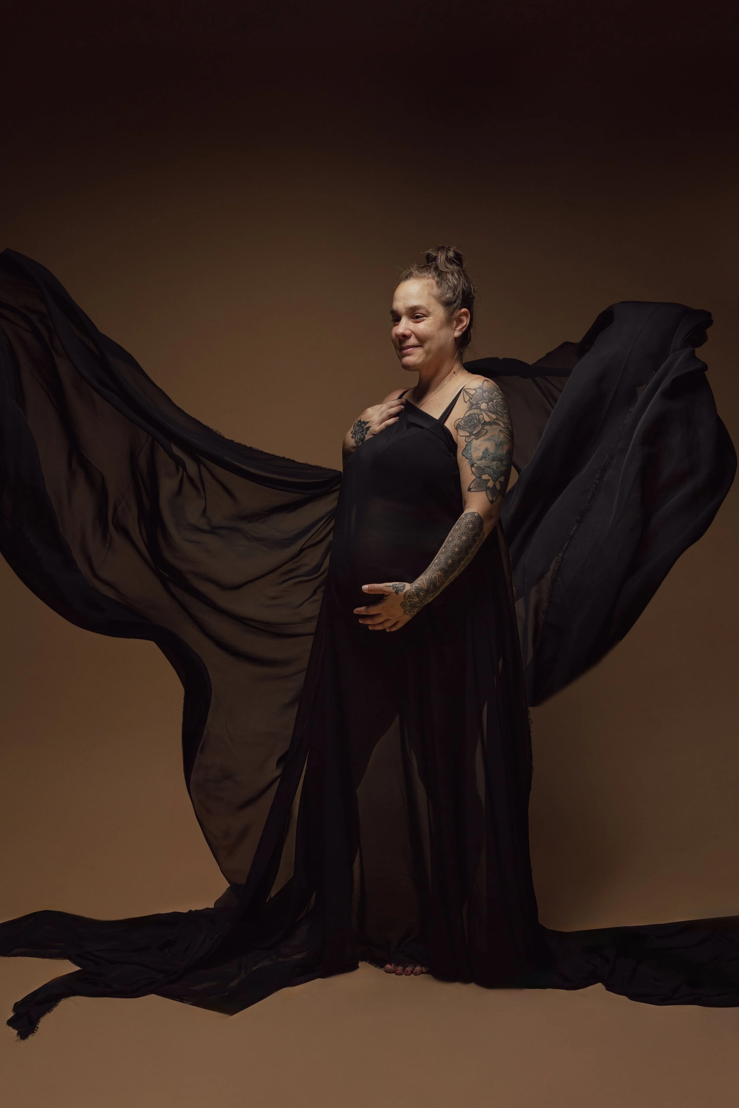 Een vrouw in een zwarte jurk poseert met grote zwarte stoffen vleugels tegen een effen bruin achtergrond.