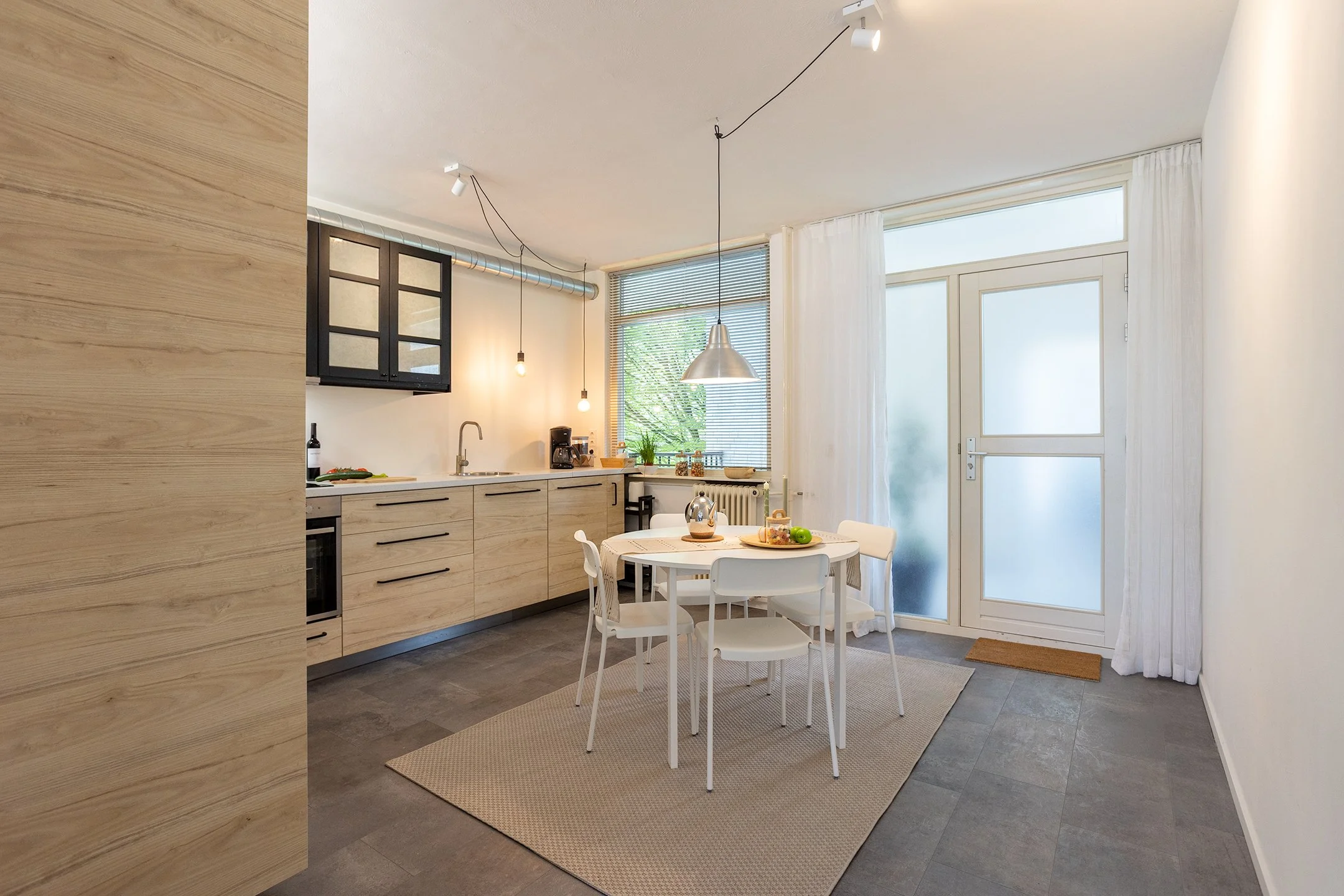 Een moderne keuken en eetkamer met licht houten kasten, een ronde eettafel met witte stoelen, en grote ramen die daglicht binnenlaten.