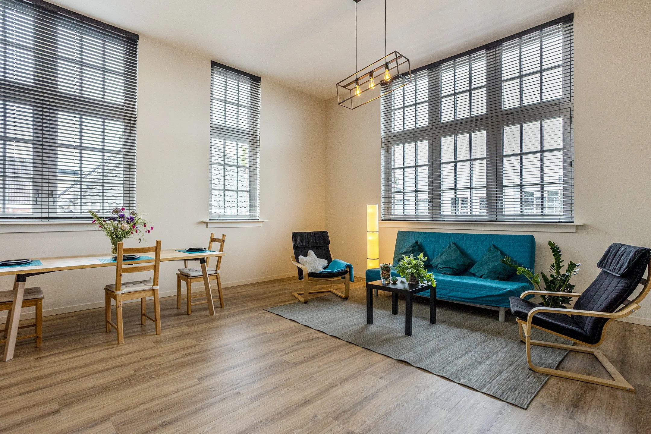 Woonkamer met groot raam, banken, stoelen, een salontafel, planten, en een vloerlamp.