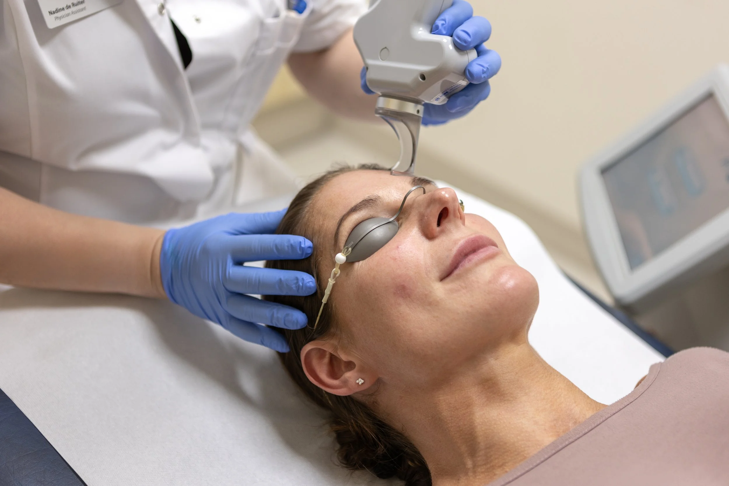 Een vrouw krijgt een medisch onderzoek met een apparaat op haar voorhoofd en draagt oogbeschermers, terwijl een arts haar helpt en een monitor zichtbaar is op de achtergrond.