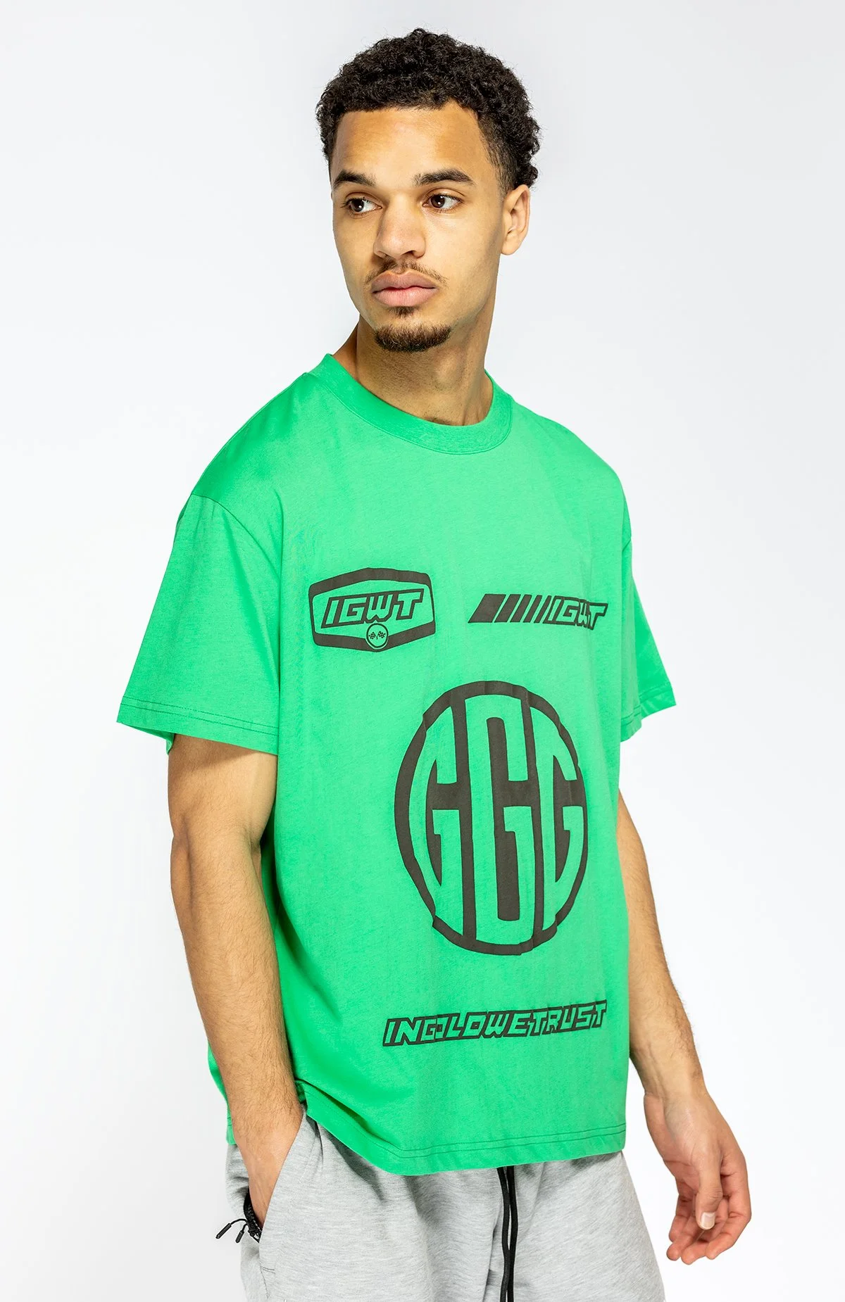 Man draagt een groene T-shirt met zwarte grafische ontwerpen en teksten, korte mouwen, en grijze sweatpants.