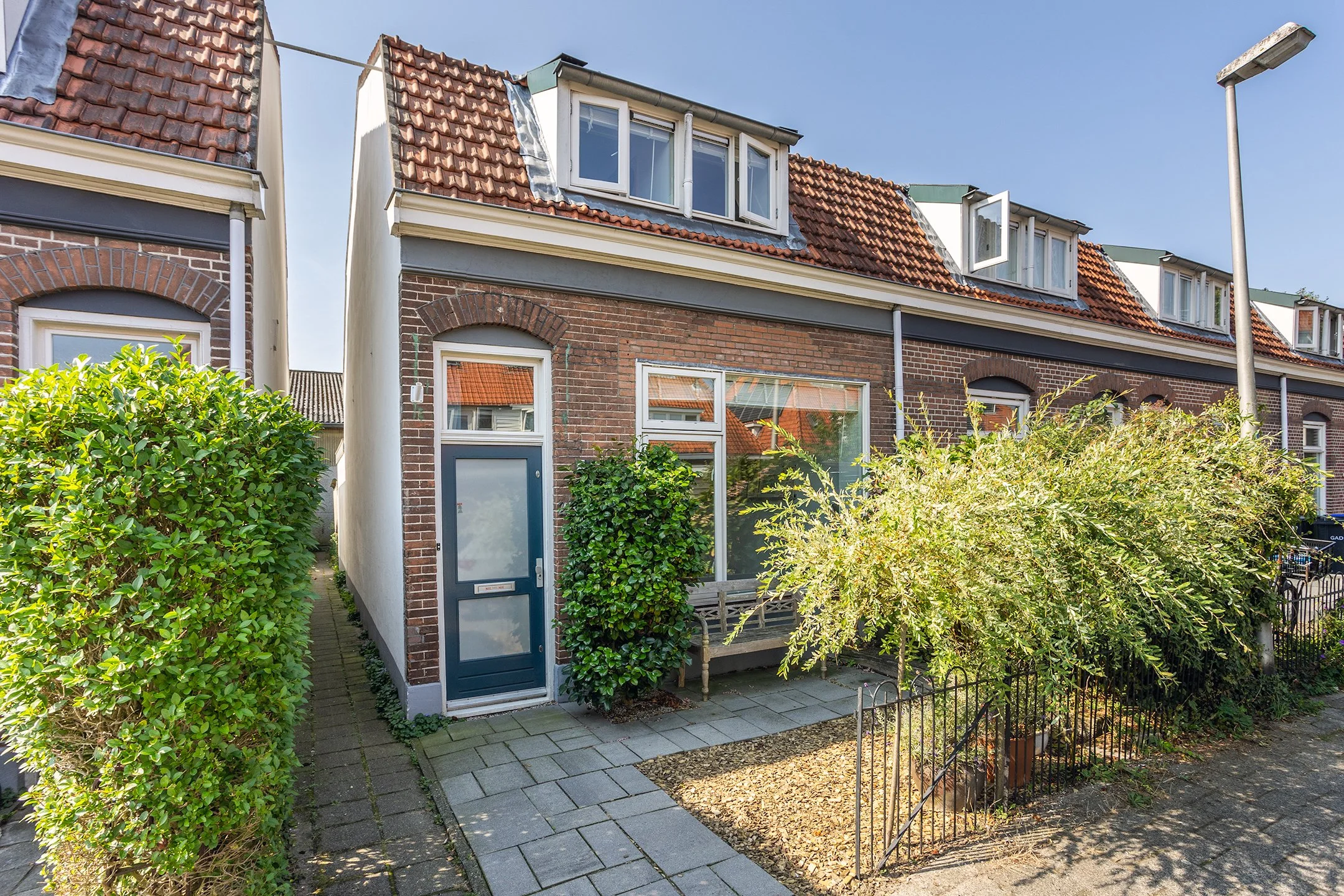 Rijtjeshuis met rode dakpannen, glasdeur, groene struiken en een bankje voor de ingang, onder een heldere blauwe hemel.