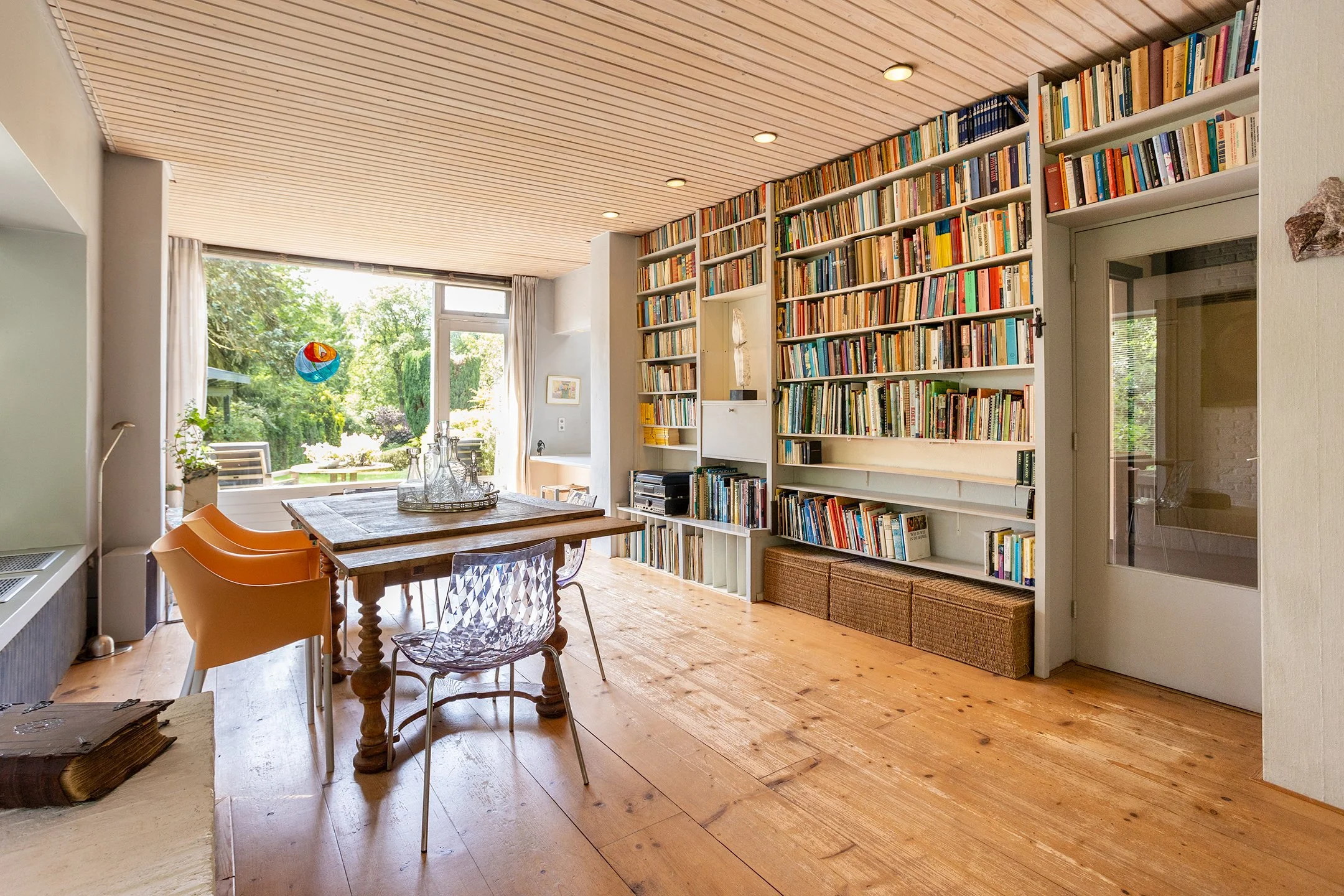 Interieur van een woonkamer of studeerkamer met grote boekenkast, houten vloer, houten plafond, zithoek bij het raam, en een eettafel met diverse stoelen.